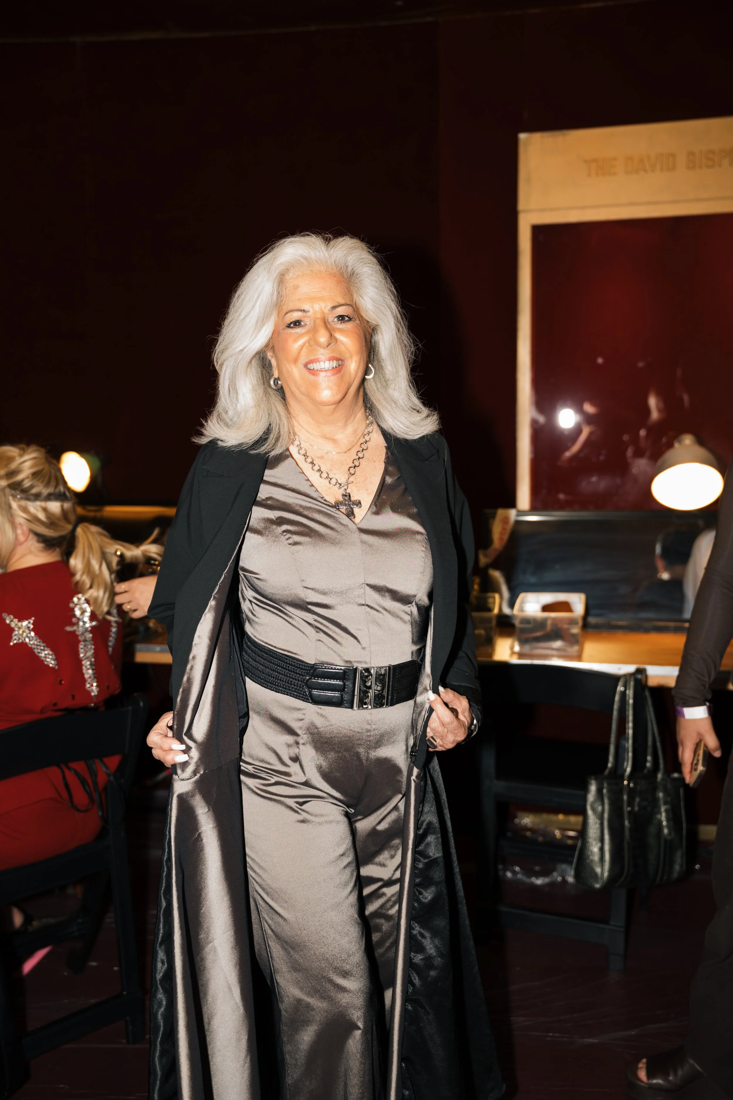 An elderly woman with gray hair smiling, dressed in a shiny gray jumpsuit with a black belt and a black coat, standing indoors at a social event with other people in the background.