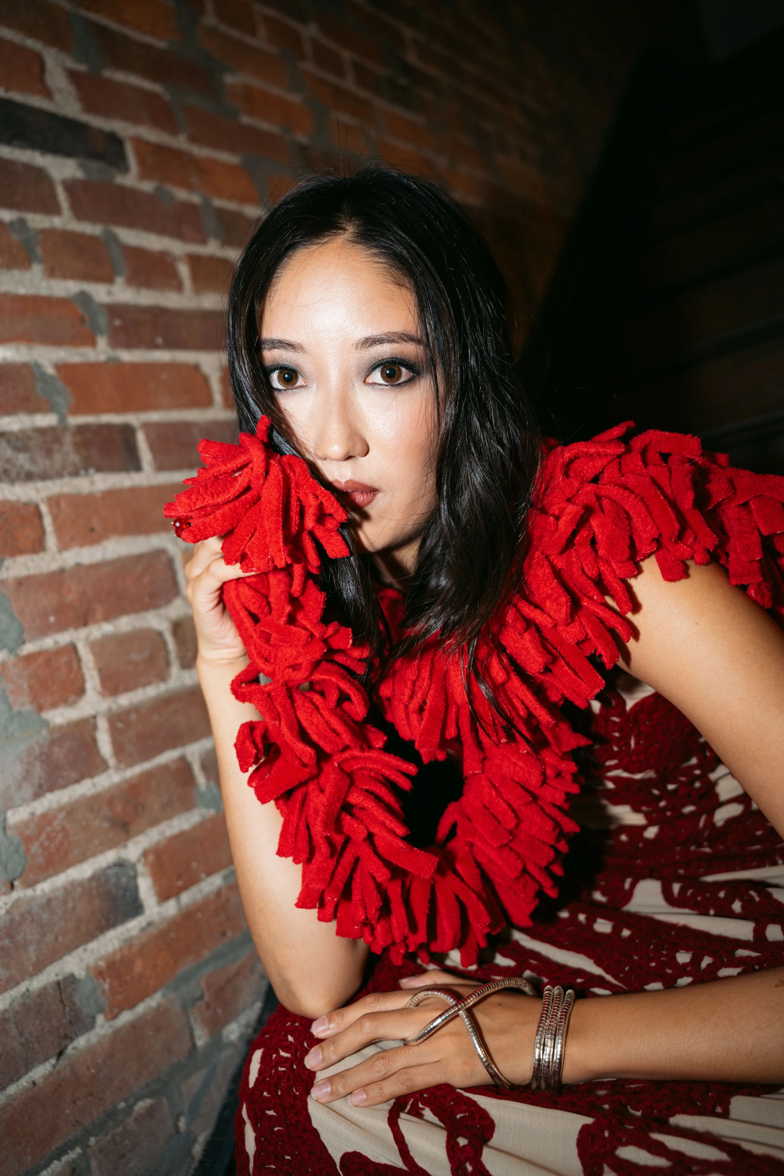 A woman with long dark hair and brown eyes is wearing a red, textured scarf and a sleeveless top. She is sitting against a brick wall and resting her arm on her lap, holding a small purse and wearing multiple bracelets.