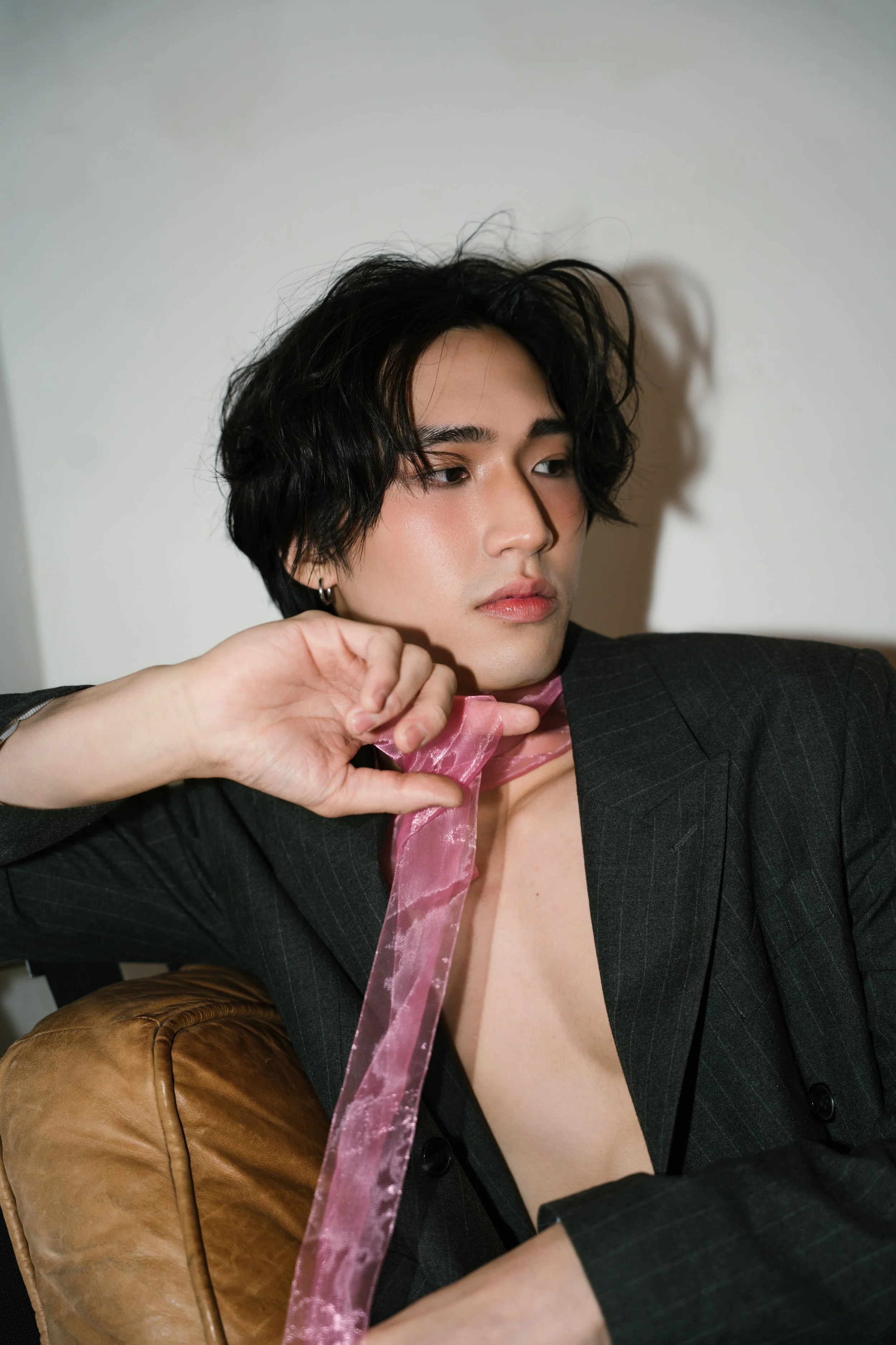 A person with short wavy black hair, wearing a dark blazer with a pink ribbon tied around their neck, sitting on a brown chair against a plain white wall.