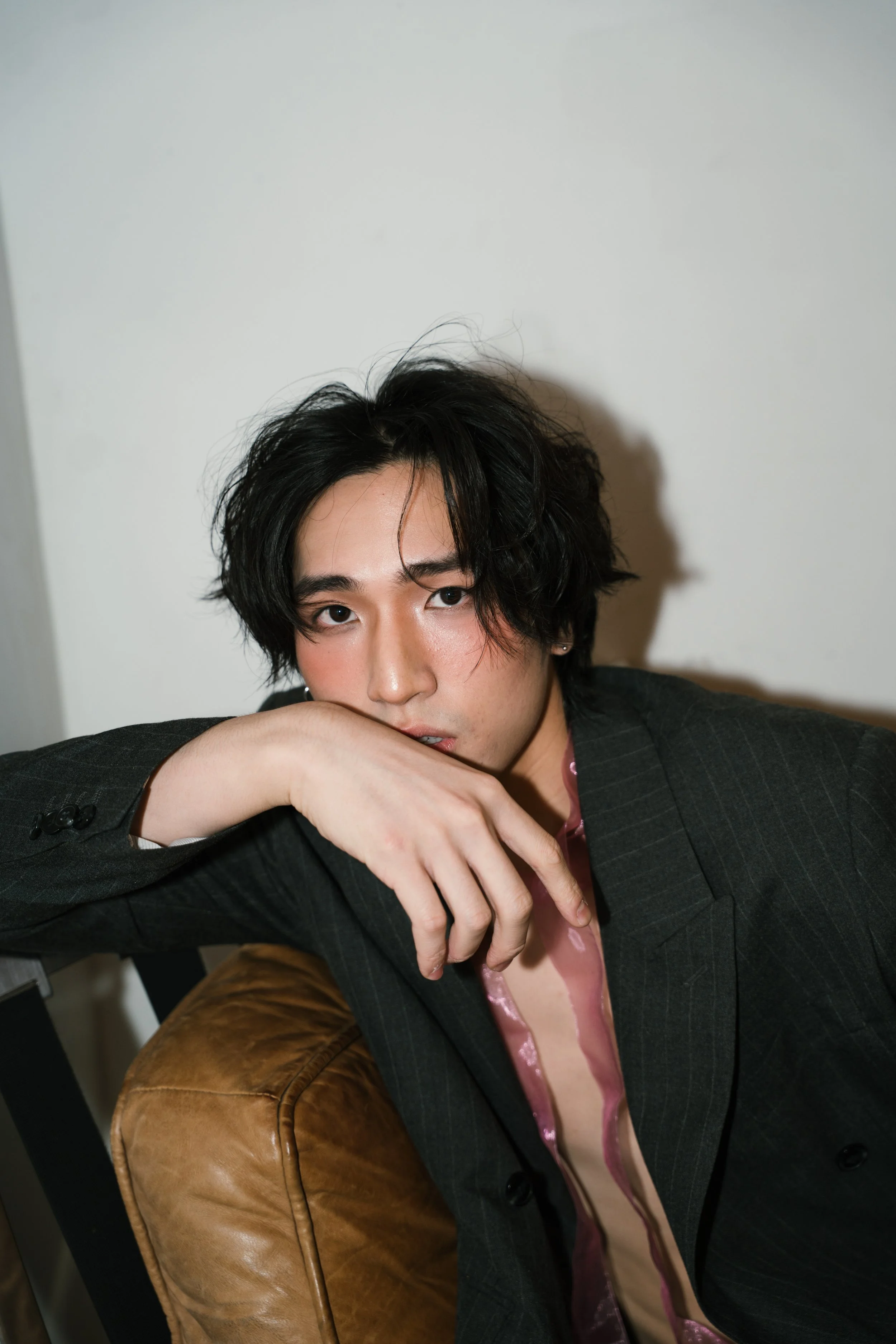 A young man with black, wavy hair, wearing a dark pinstripe suit jacket over a shiny pink shirt, sitting on a brown leather chair. He is resting his face on his arm and looking at the camera.