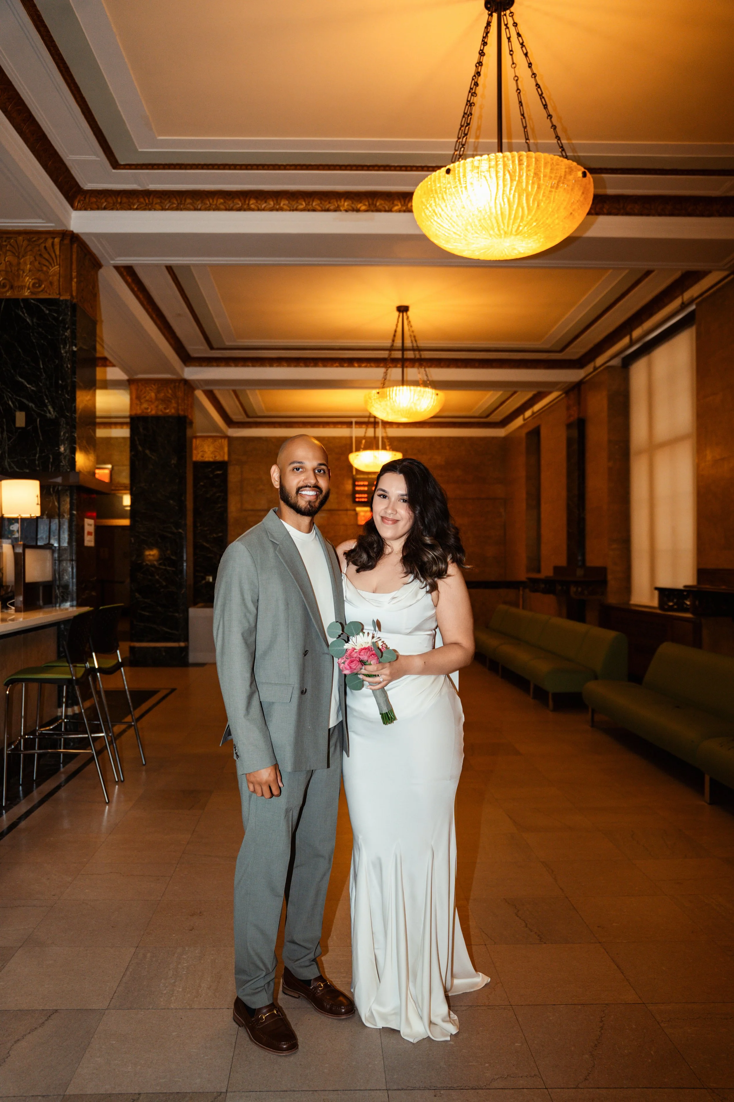 A man and woman dressed in formal attire, standing together in an elegant, warmly lit lobby. The woman holds a bouquet of pink flowers.