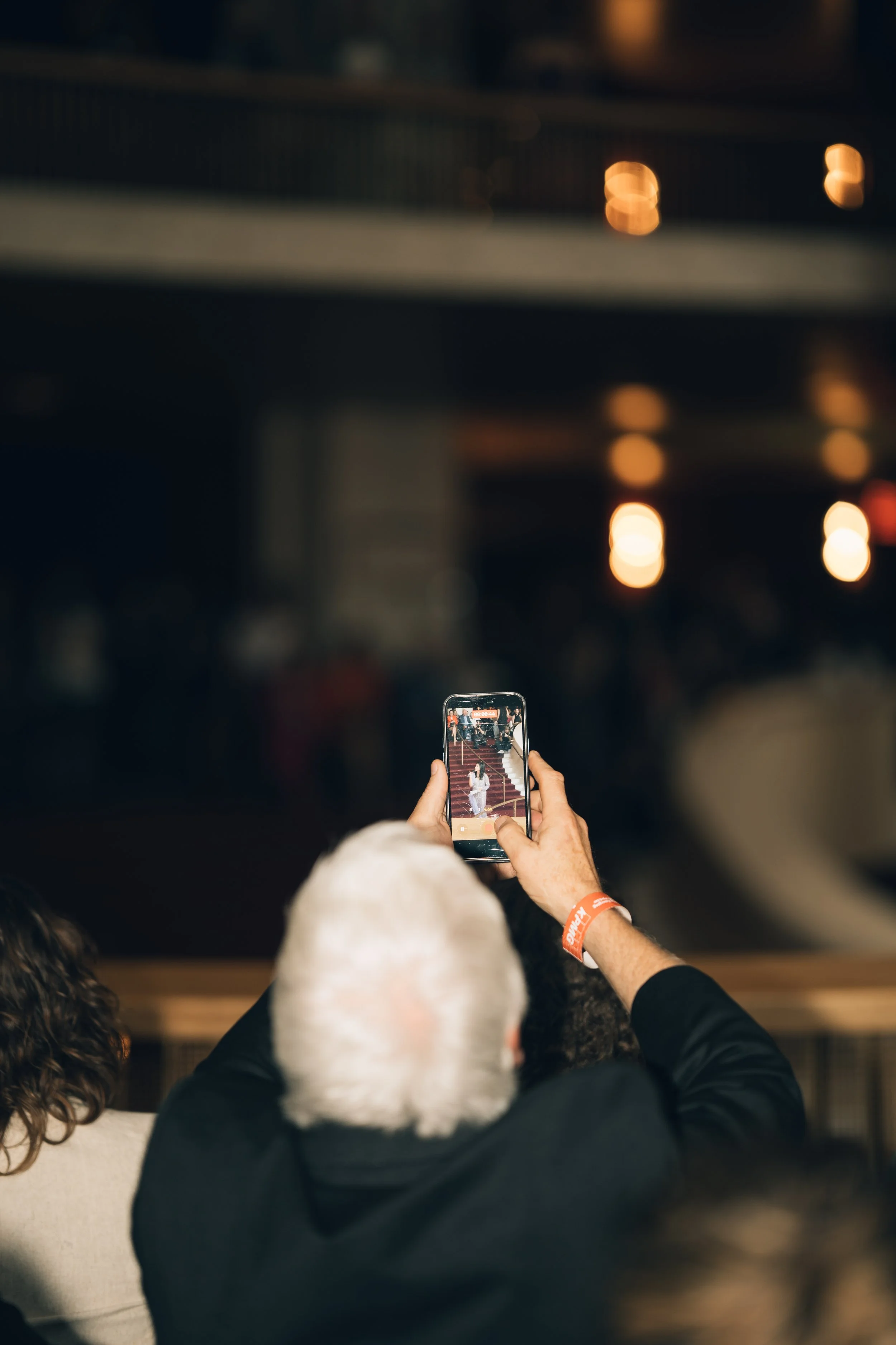 Person taking a photo of a performer on stage with a smartphone in a dimly lit auditorium.