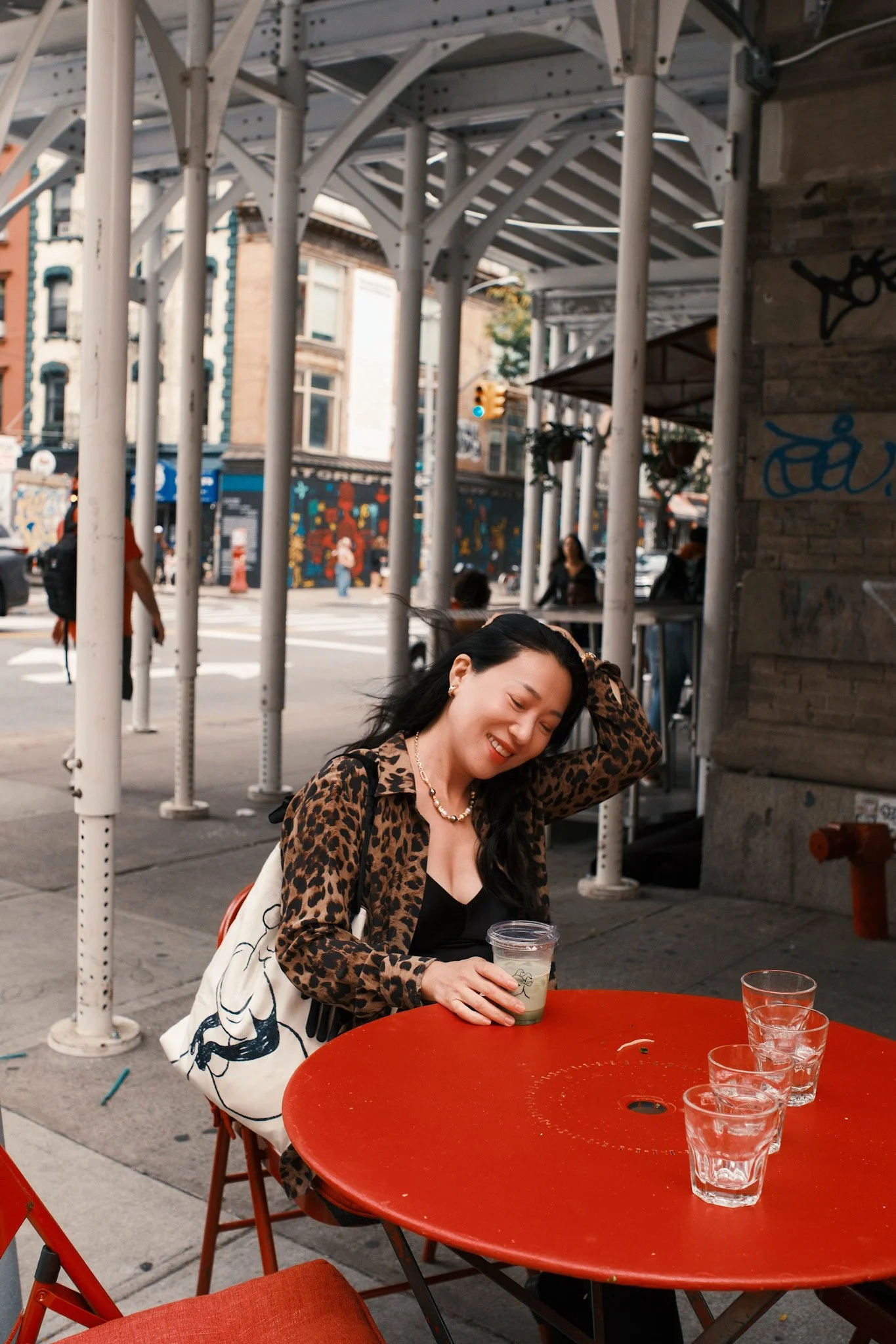 A woman with black hair smiling and sitting at a red outdoor table, holding a drink in a clear plastic cup, with a city street background and graffiti on a building wall.