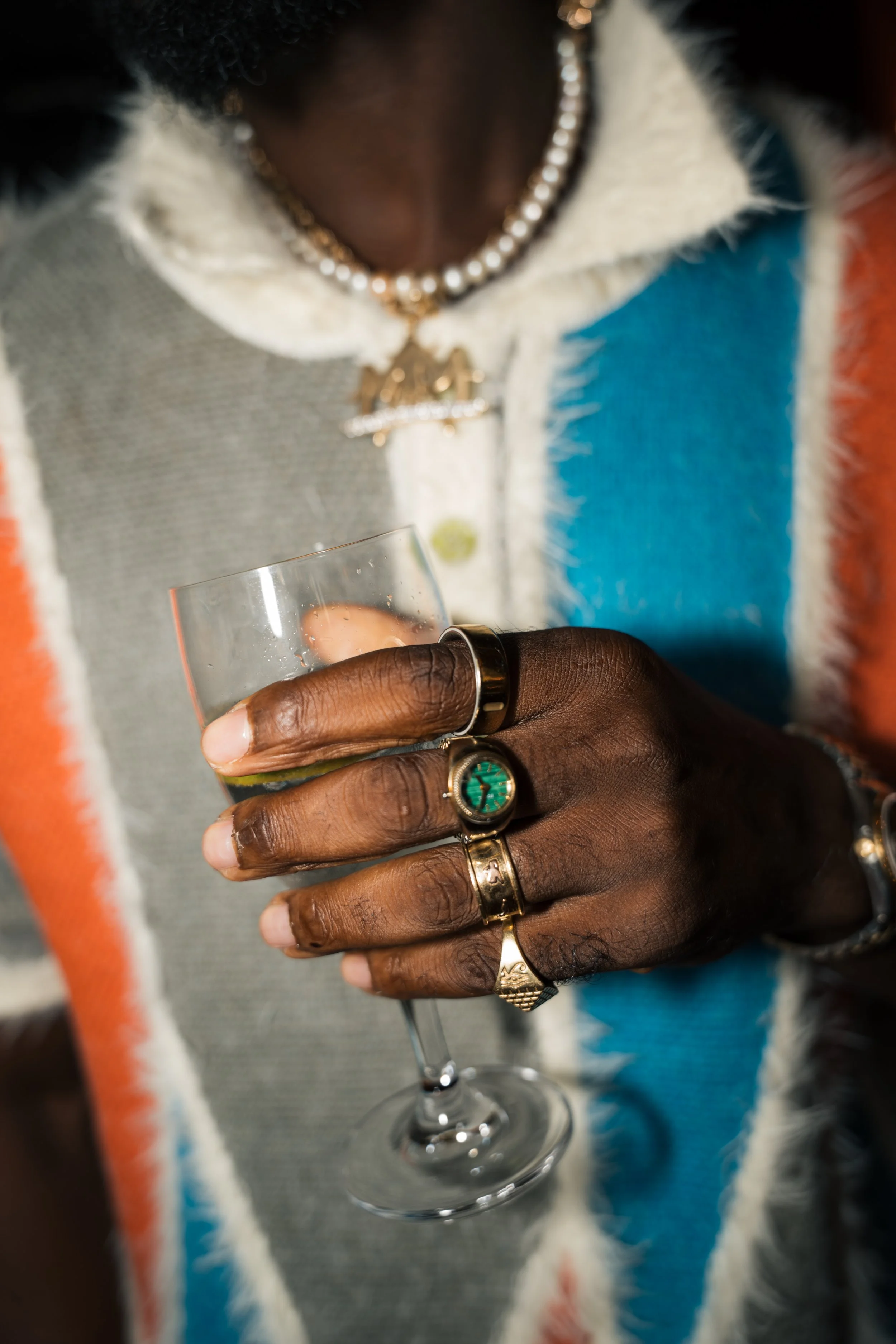 Close-up of a person's hand holding a glass of champagne. The person is wearing multiple rings, necklaces, and a colorful, textured jacket with white, orange, blue, and gray colors.