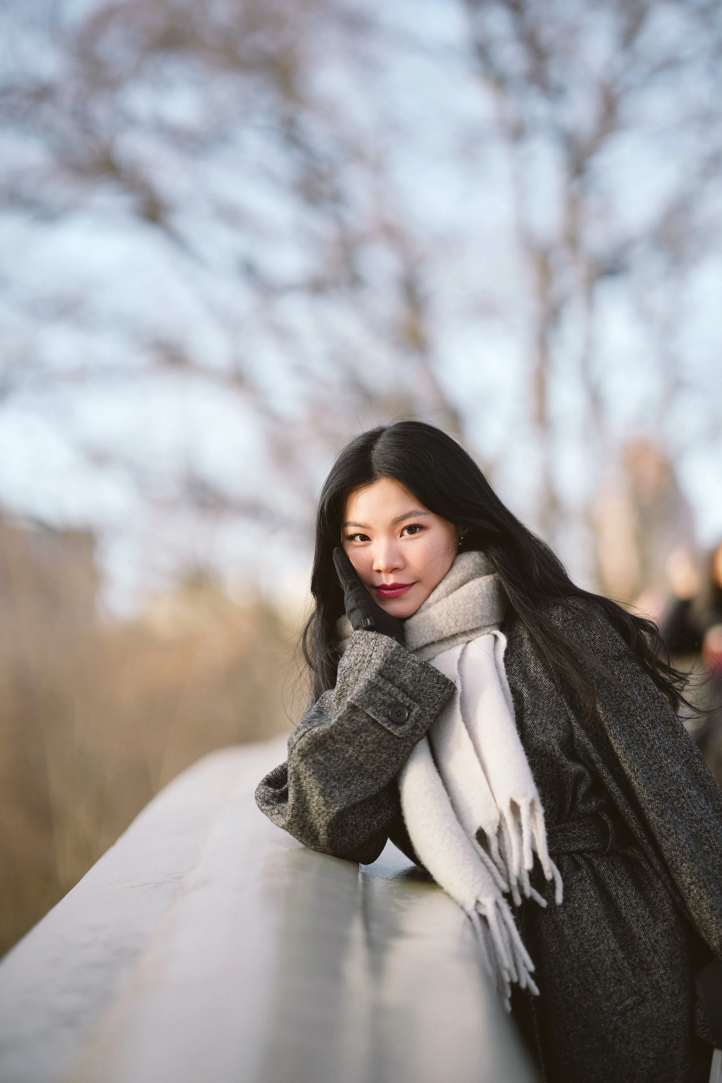 A young woman with long dark hair leaning on a white railing outdoors during winter. She is dressed in a gray coat, white scarf, and black gloves. Snow-covered trees are blurred in the background.