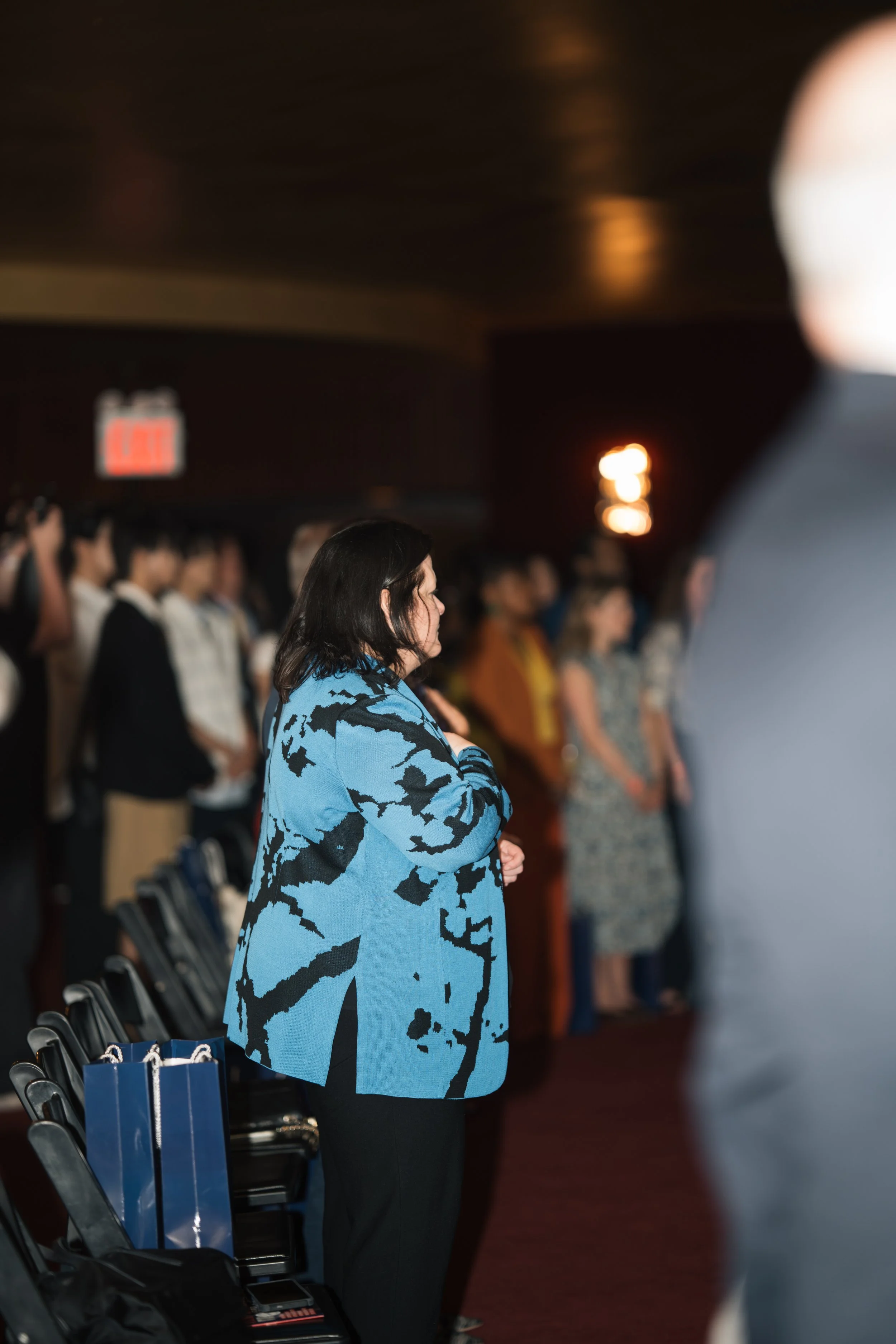 A woman with dark hair wearing a blue and black patterned coat, standing with her hands clasped, appears to be praying or reflecting during a formal event in a dimly lit auditorium filled with other attendees.