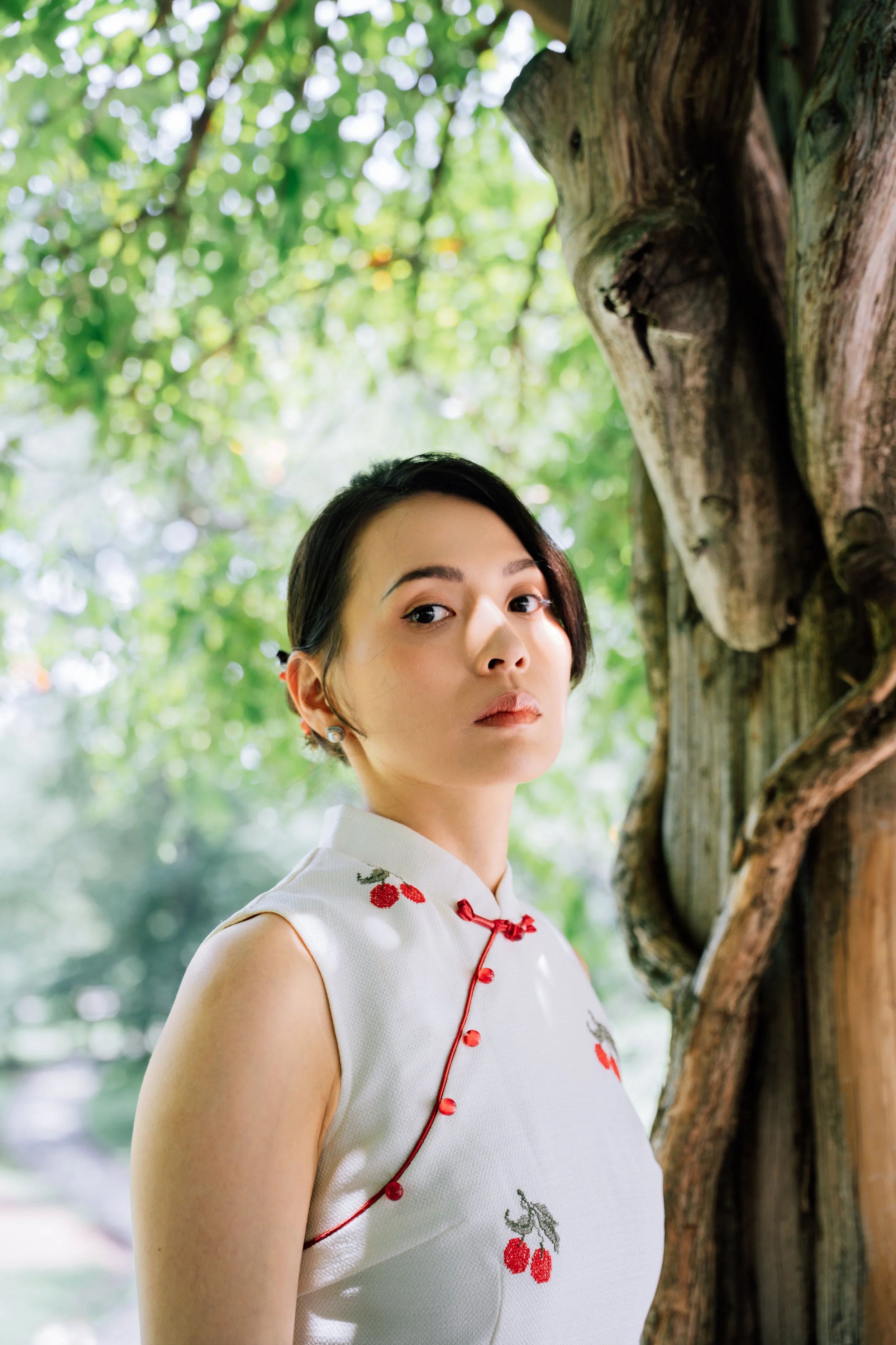 A woman in a white, cherry-embroidered traditional Chinese dress standing outdoors next to a tree with green foliage in the background.