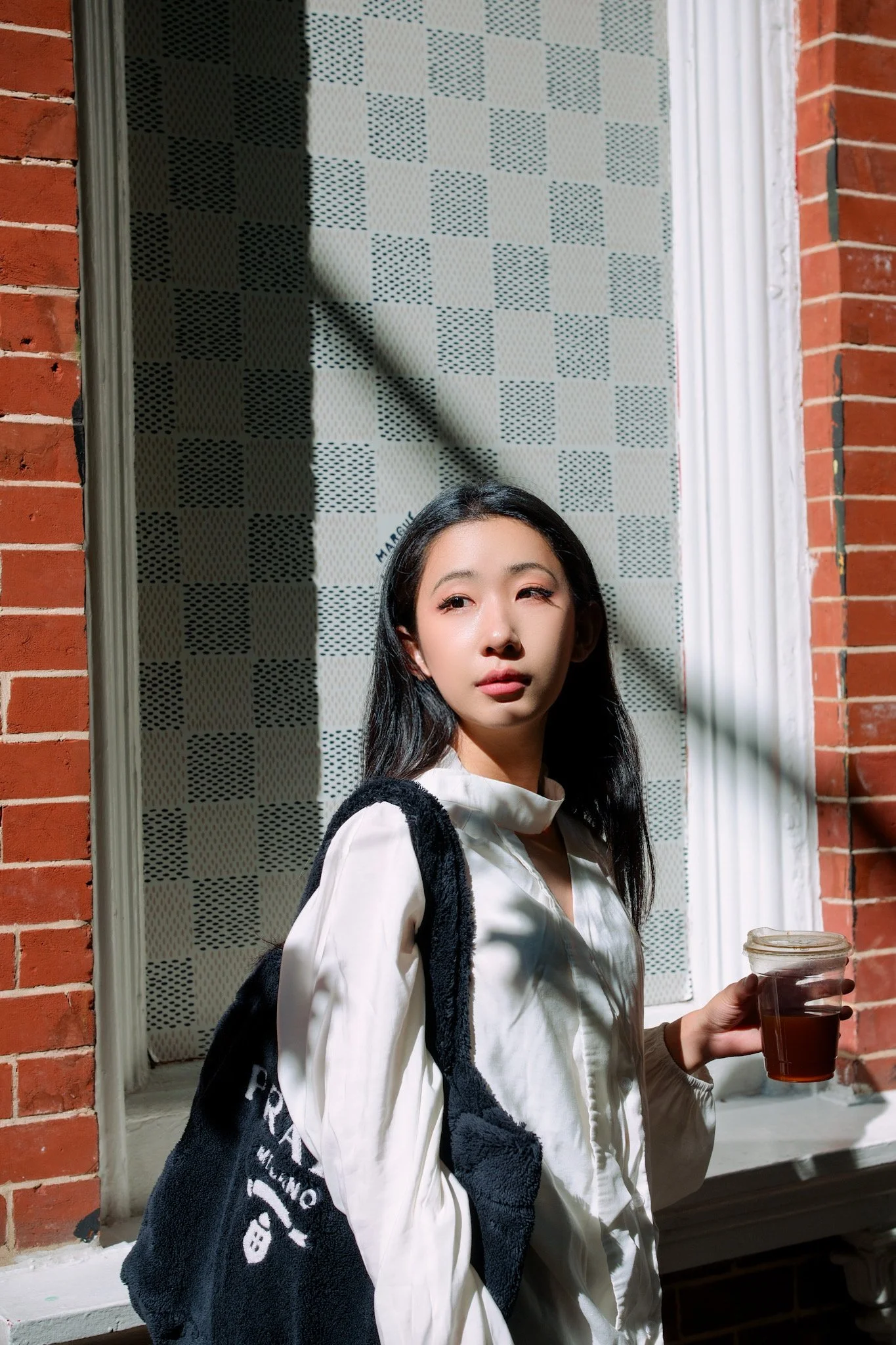 A young woman with black hair, wearing a white shirt, standing outside at night, holding a drink in her right hand, with a brick wall and window in the background.