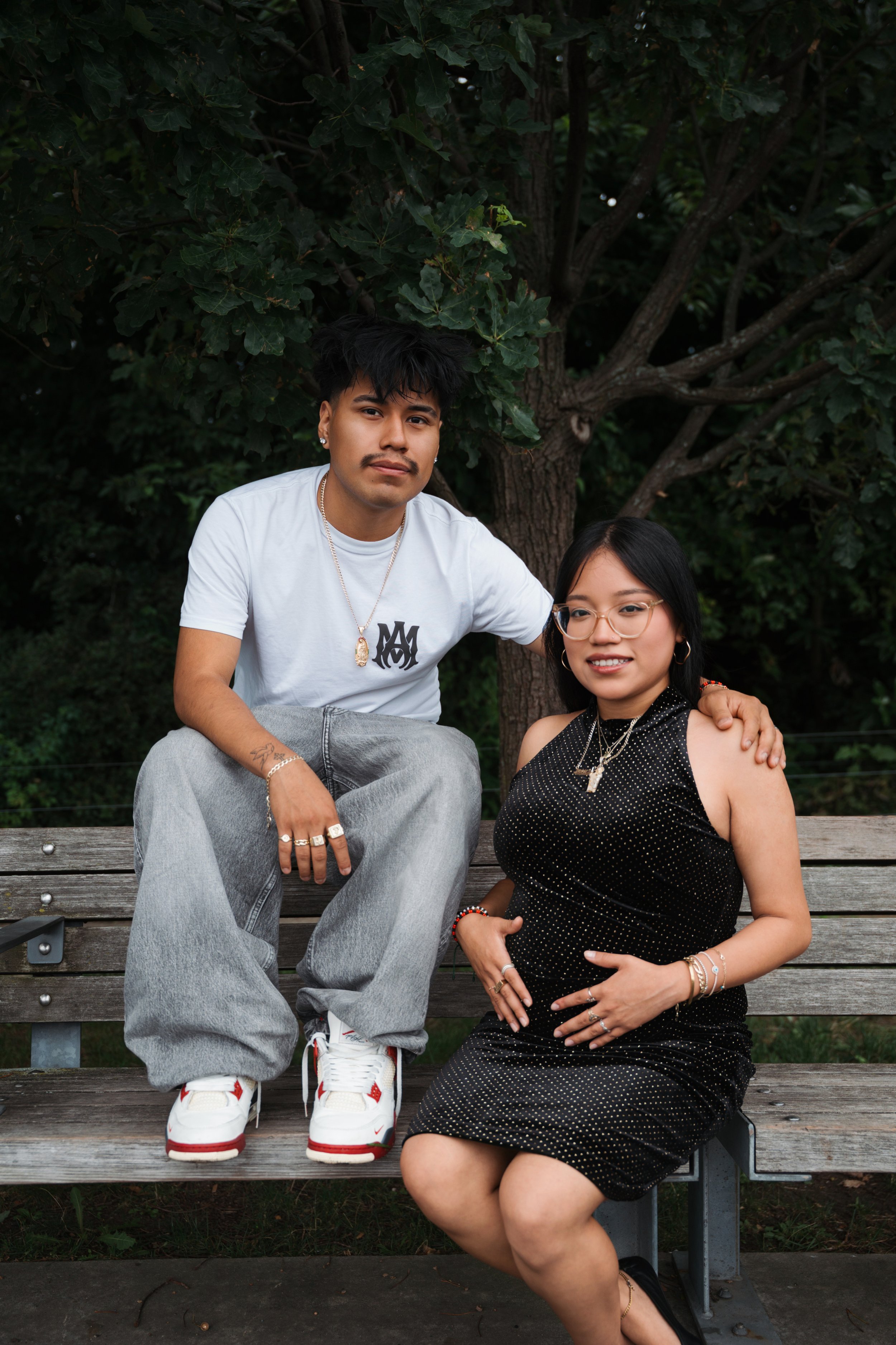 A young man and woman sitting on a park bench with a tree and greenery in the background. The man has black hair, facial hair, tattoos, and is wearing a white t-shirt, grey jeans, and white sneakers. The woman has black hair, glasses, and is wearing 