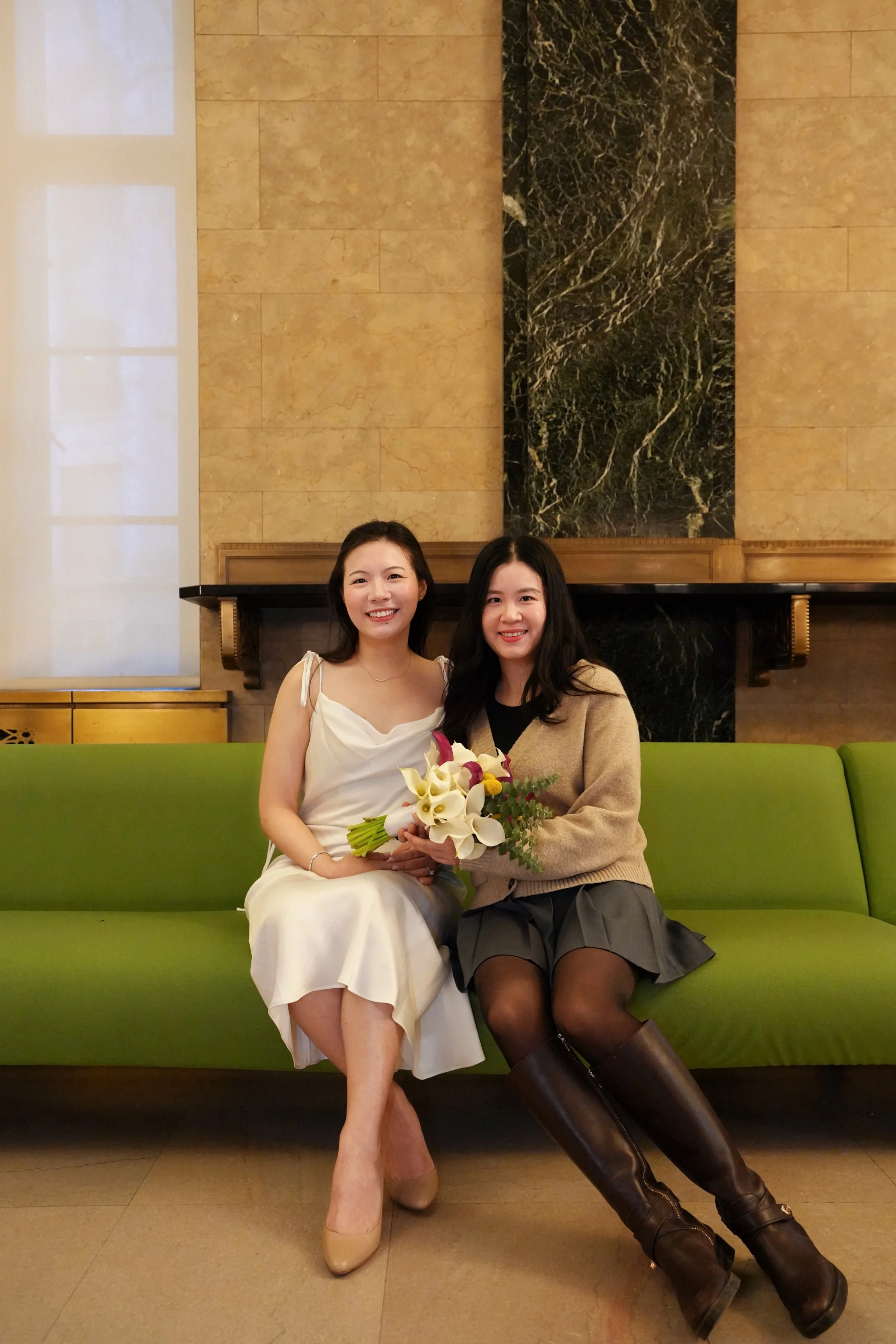 Two women sitting on a green bench in a room with beige and black marble wall background. One woman is in a white dress holding a bouquet of flowers, and the other is in a beige cardigan and gray skirt wearing black stockings and brown knee-high boot