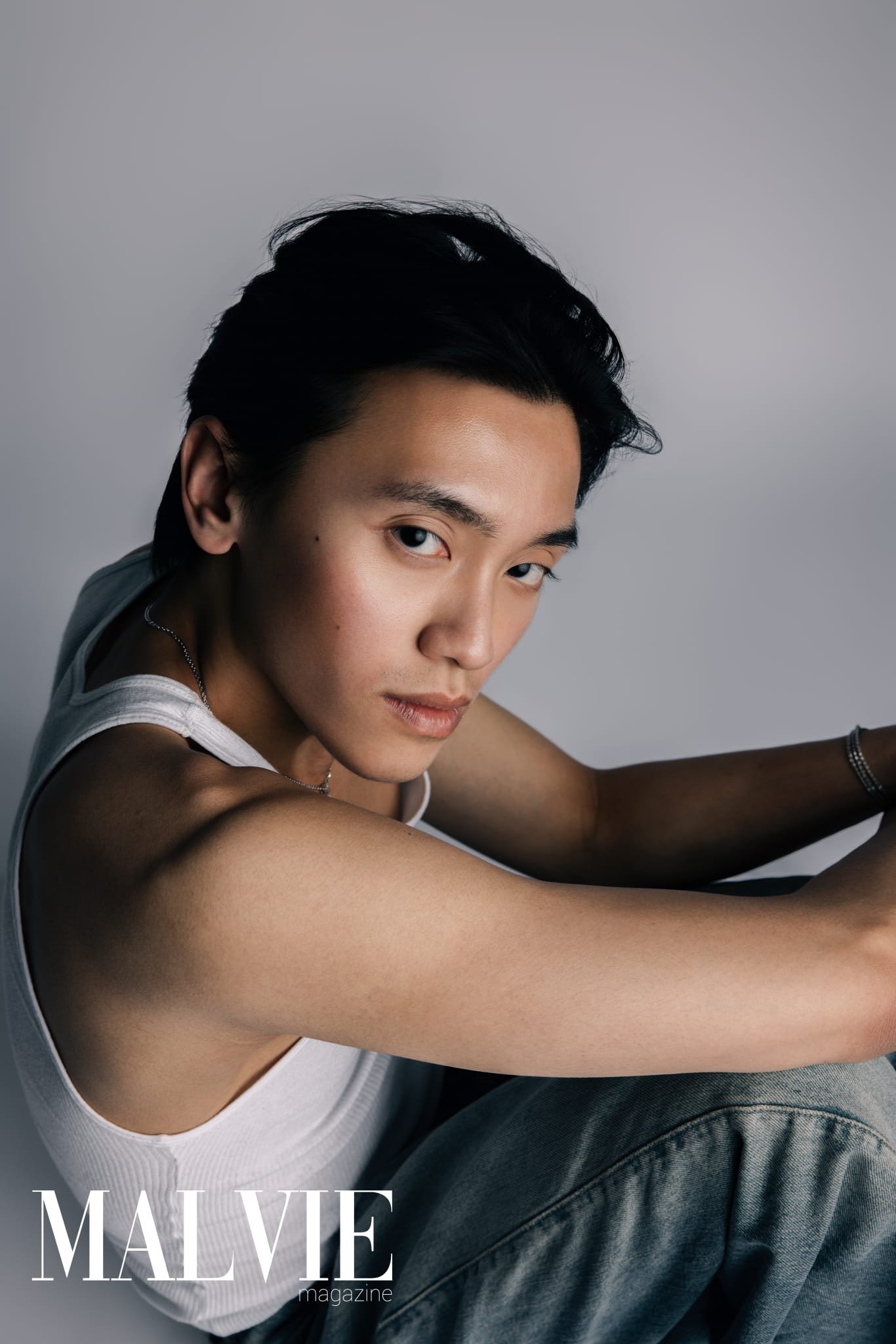 Close-up of a young man with black hair and light skin, wearing a white tank top and a silver necklace, leaning forward with his arms stretched out, against a plain gray background.