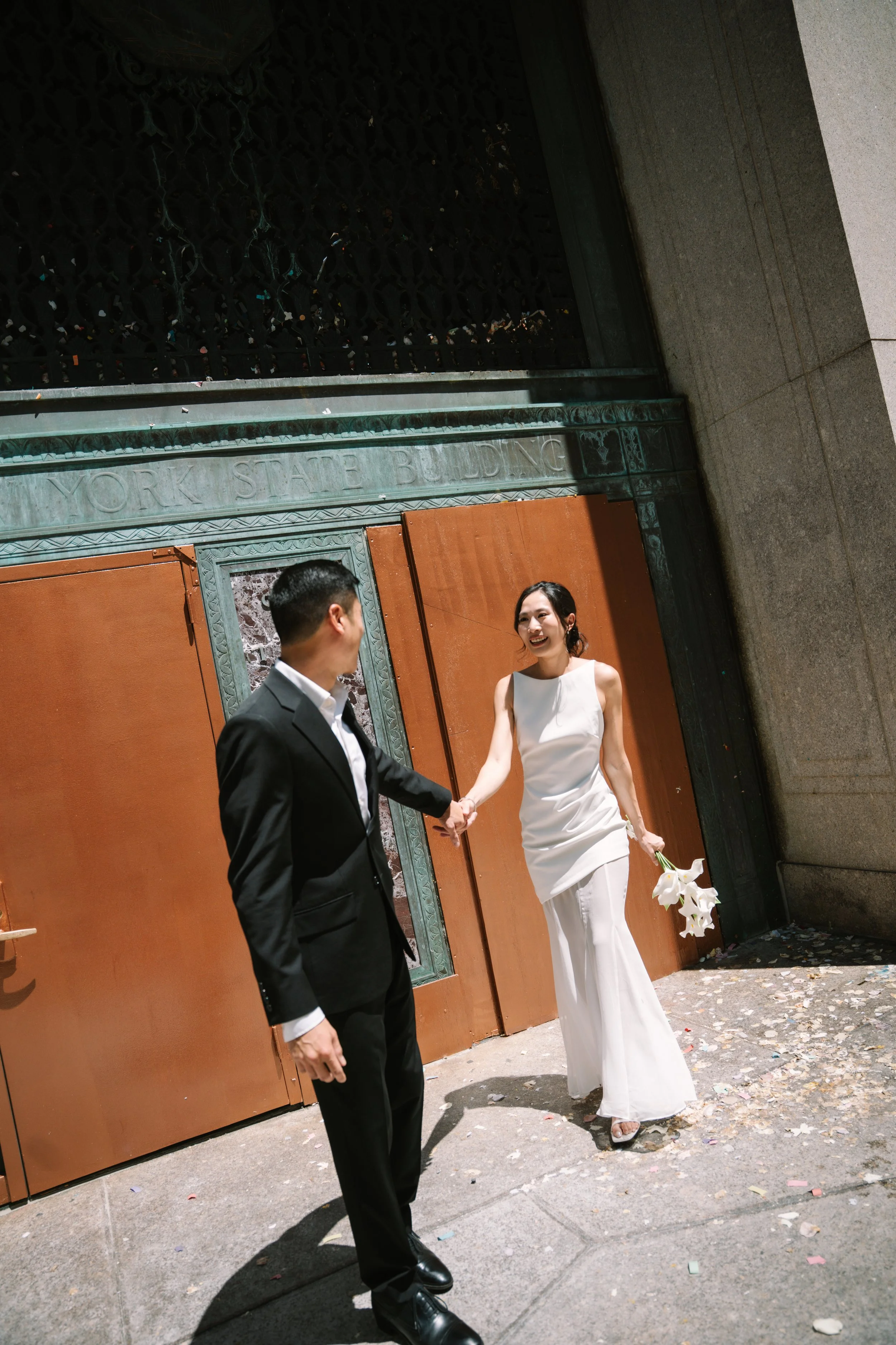 A couple dressed in wedding attire holding hands outside a building entrance labeled 'YORK STATE BUILDING.' The woman is in a white dress holding white flowers, and the man is in a black suit. They are smiling and looking at each other, with confetti