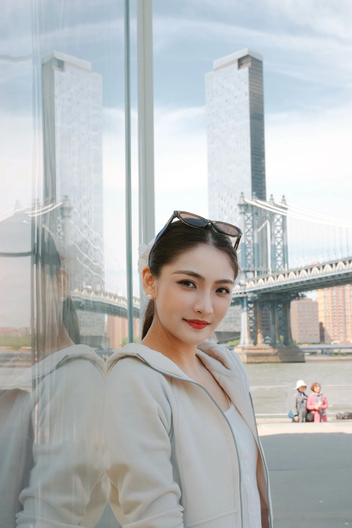 A woman with sunglasses on her head and pearl earrings, smiling in front of a glass panel with a city bridge and tall buildings in the background.