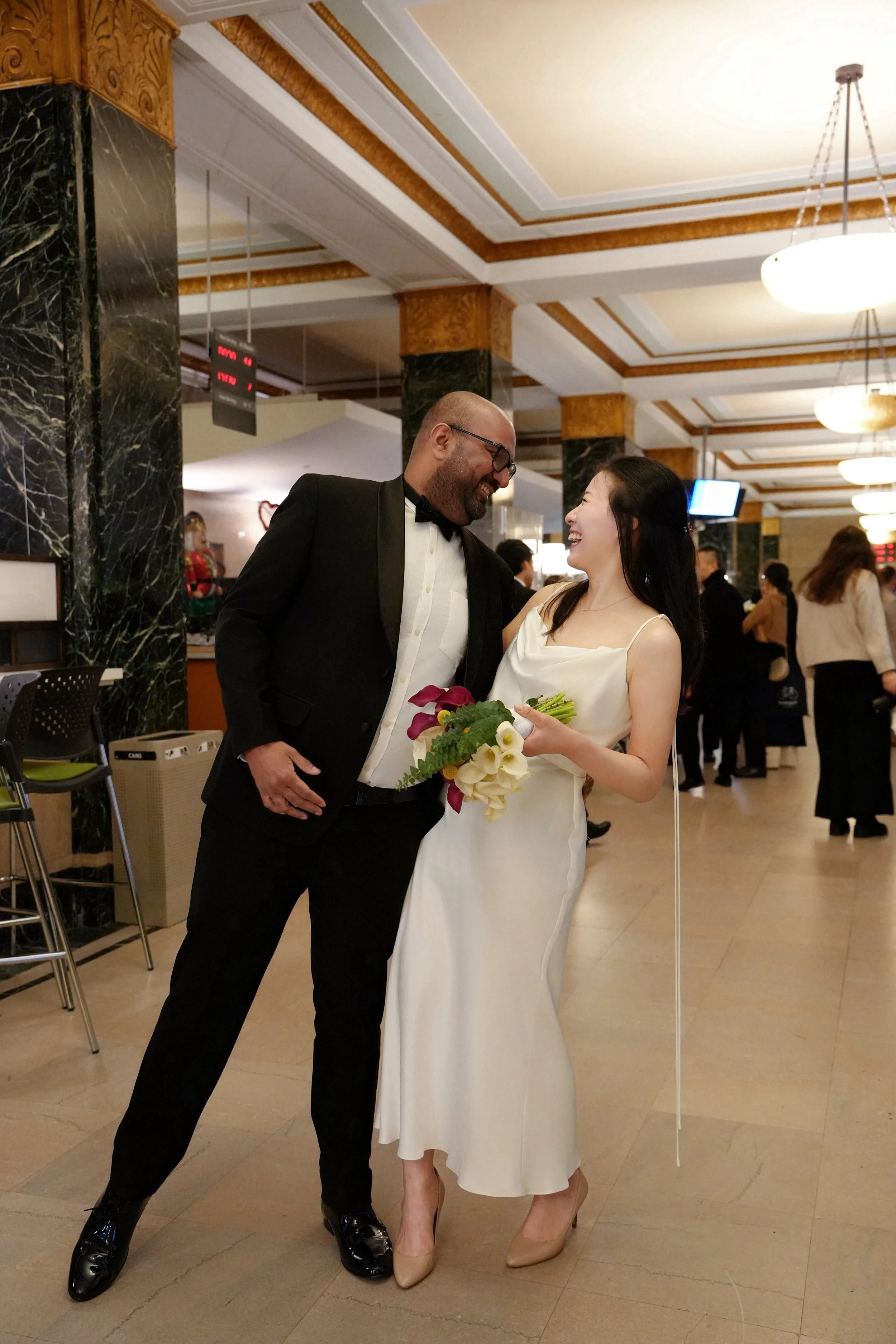 A man in a tuxedo and a woman in an elegant white dress are smiling and interacting at a formal event. The woman is holding a bouquet of flowers. The background shows a decorated ceiling and other attendees in formal attire.