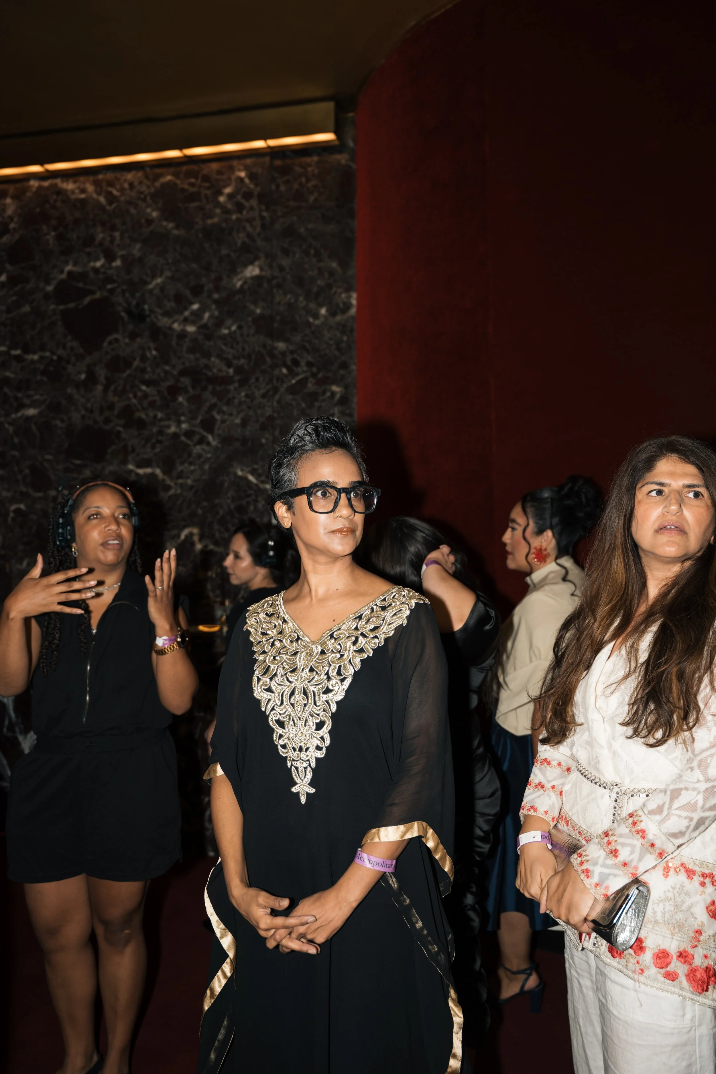 A group of women at an indoor event. The woman in the center has short gray hair, glasses, and is wearing a black dress with ornate gold embroidery. Other women around her are talking and wearing various outfits, including headphones, and appear enga