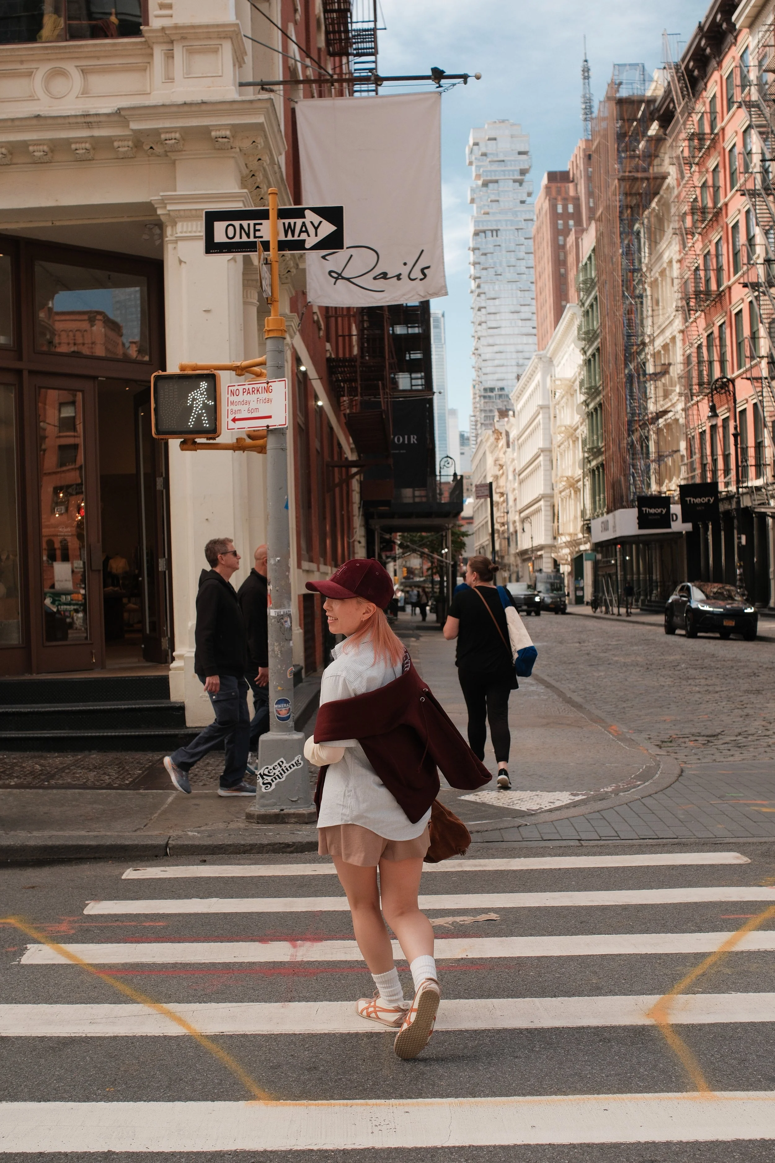A woman with pink hair and a cap crossing a city street at a crosswalk with people walking on the sidewalk and buildings in the background.