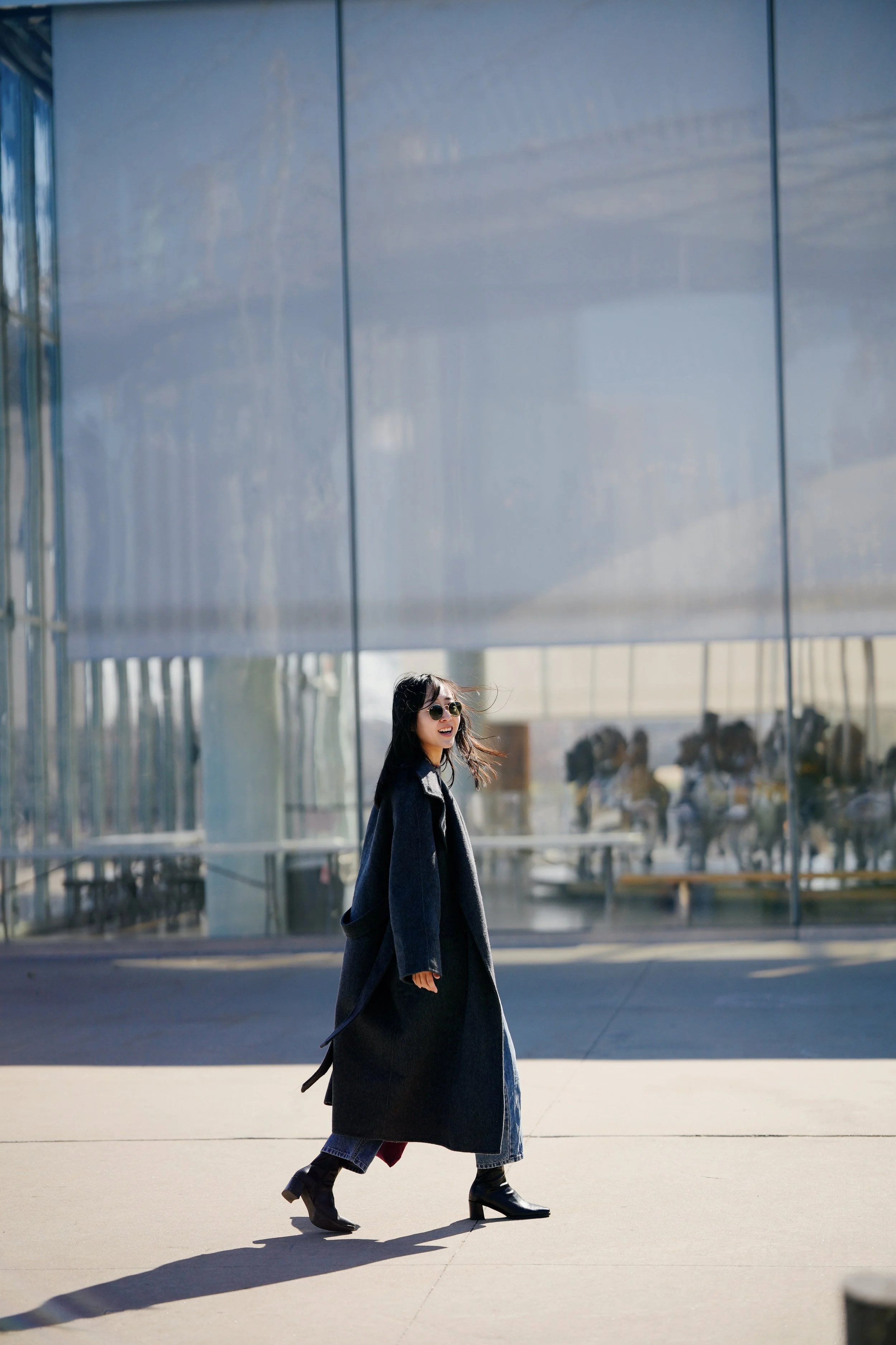 A woman walking outdoors in front of modern glass building, wearing a black long coat, sunglasses, and high-heeled boots, with her hair flowing and smiling.