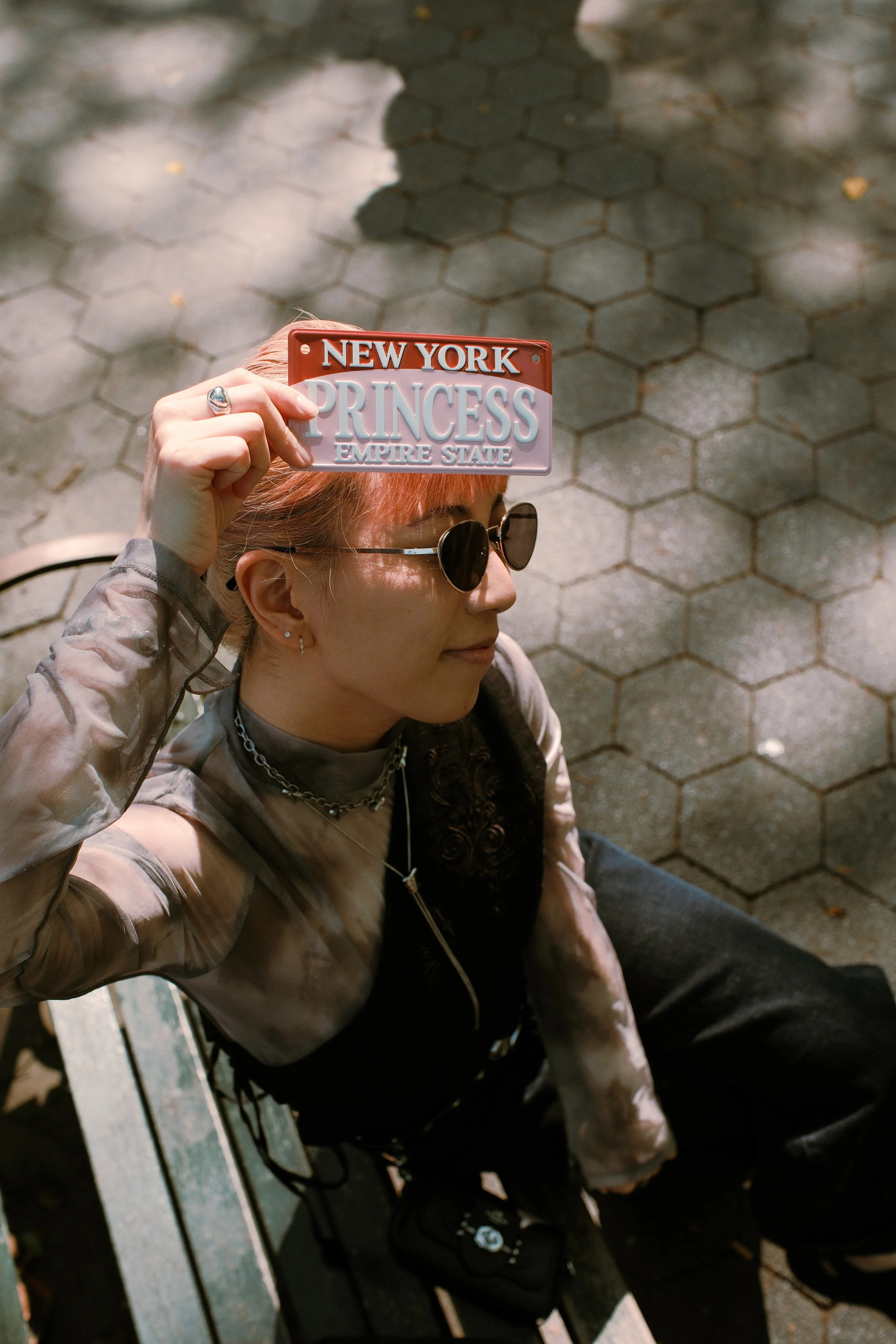 A person with light skin, short reddish hair, wearing round black sunglasses, a chain necklace, a patterned jacket, and black clothing, sitting on a park bench with a shadowed hexagonal tile ground in the background. They are holding a colorful licen