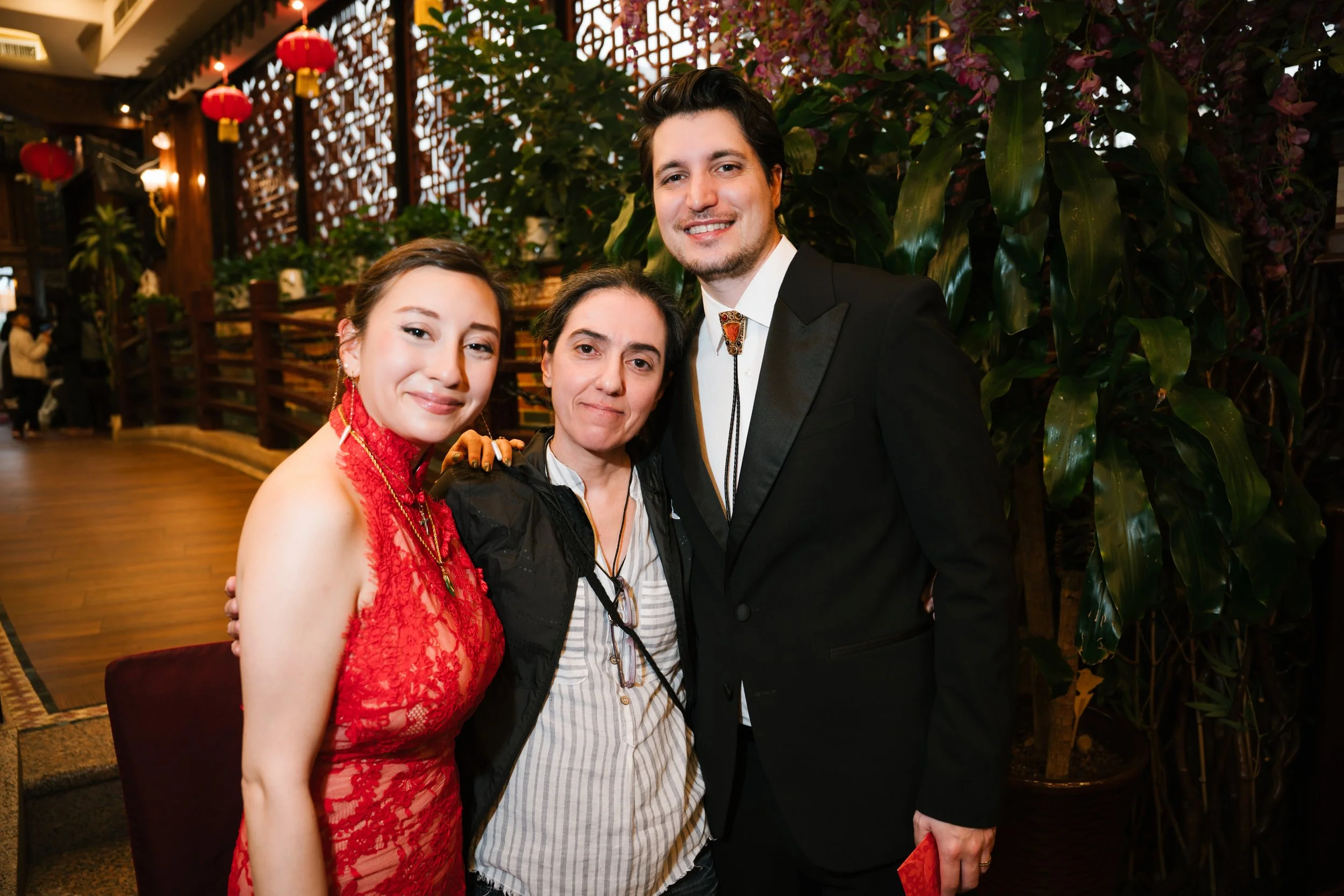 Three people posing together in a decorated indoor setting with red lanterns and lush green plants. The woman on the left is wearing a red lace dress, and the man on the right is dressed in a black tuxedo with a bolo tie. The woman in the middle is w