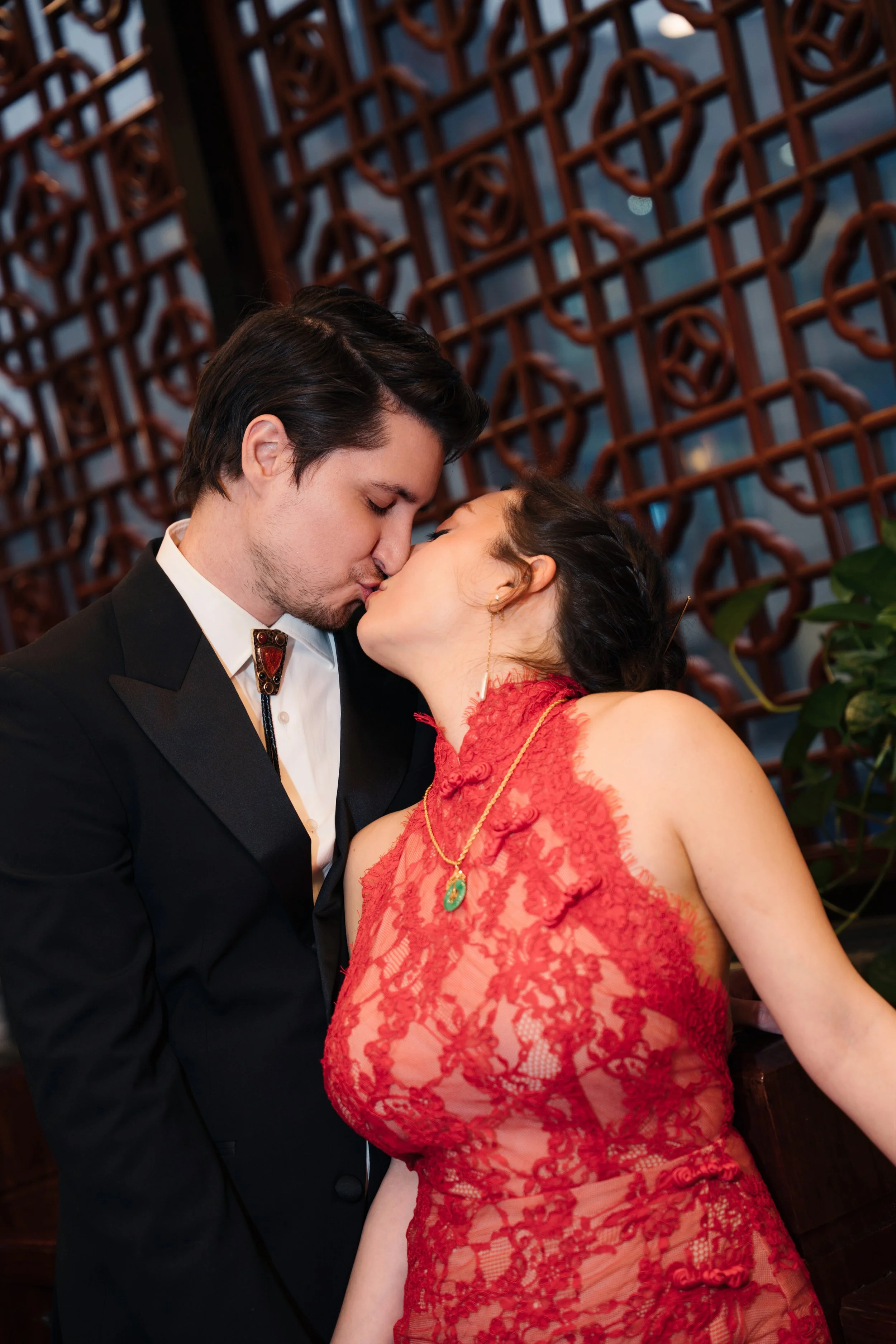 A couple kissing, the man dressed in a tuxedo and the woman in a red lace dress with a green pendant necklace, in an indoor setting with wooden lattice windows in the background.