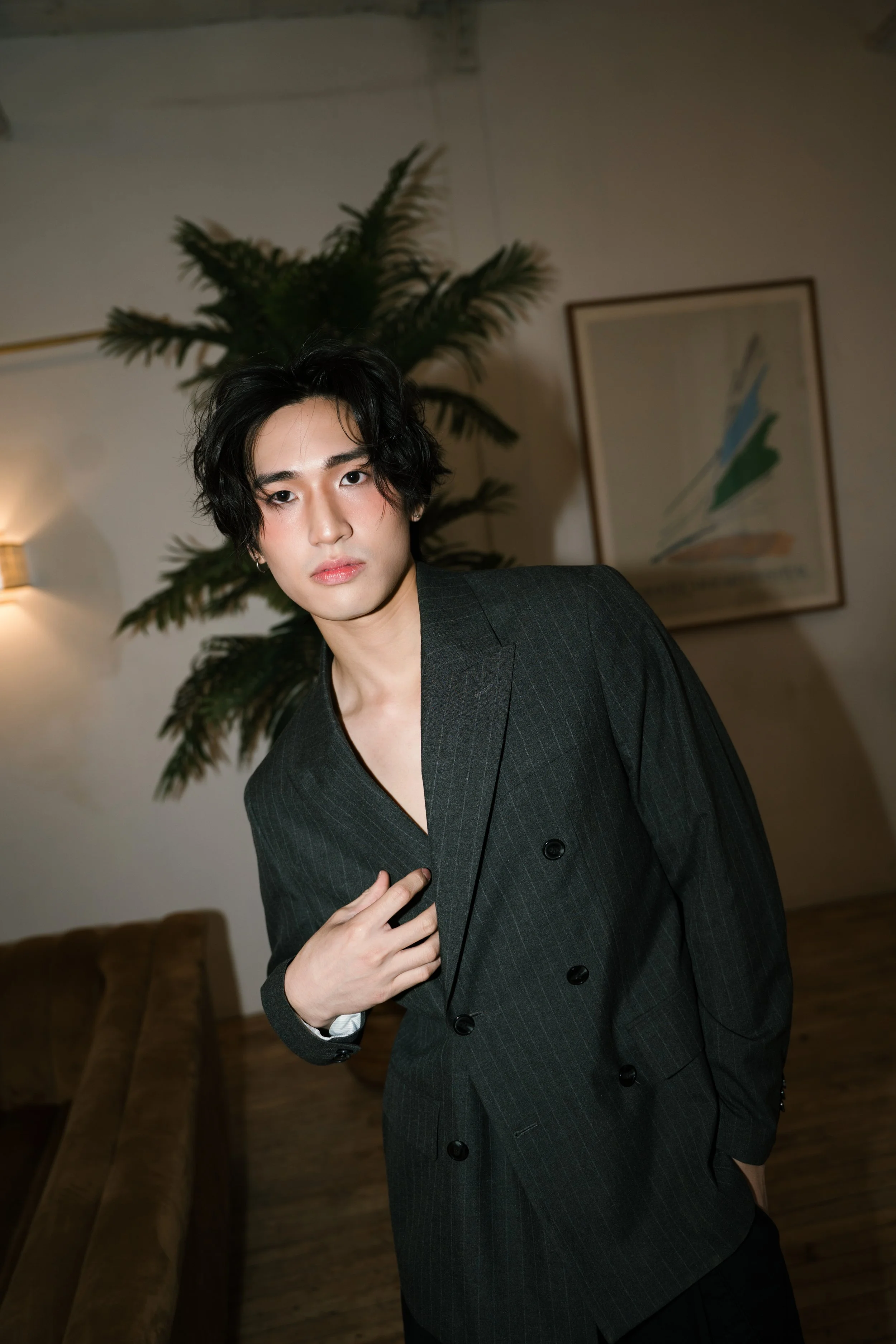 A young man with wavy black hair and fair skin wearing a dark pinstripe suit with a V-neck shirt, standing in a room decorated with a painting and a large potted plant. He is slightly leaning and touching his chest with his right hand.