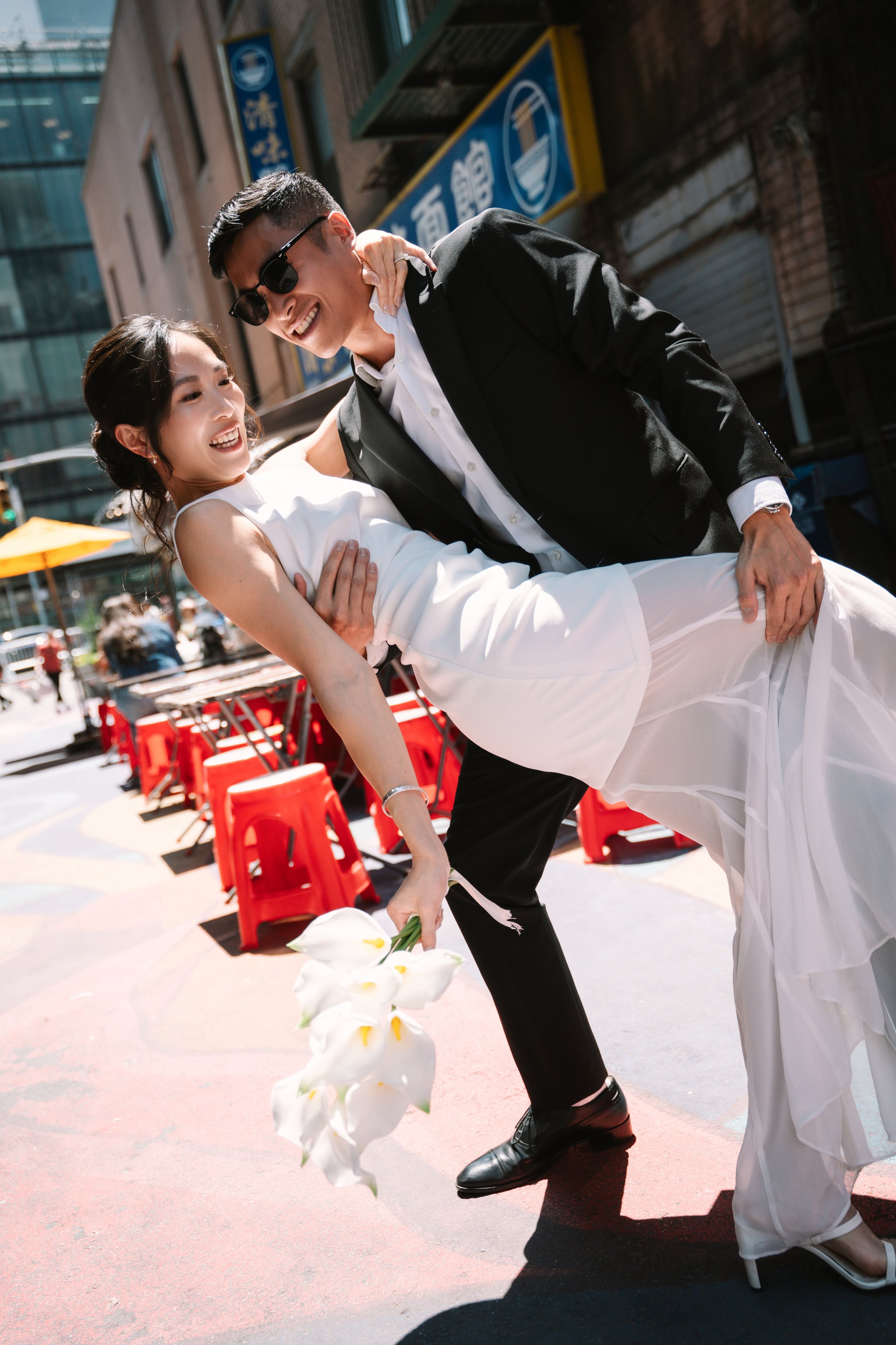 A man in a black suit and sunglasses is holding a smiling woman in a white dress. They are outdoors in an urban setting, with red chairs and a yellow umbrella in the background. The woman is holding a bouquet of white flowers, and both are happy and 
