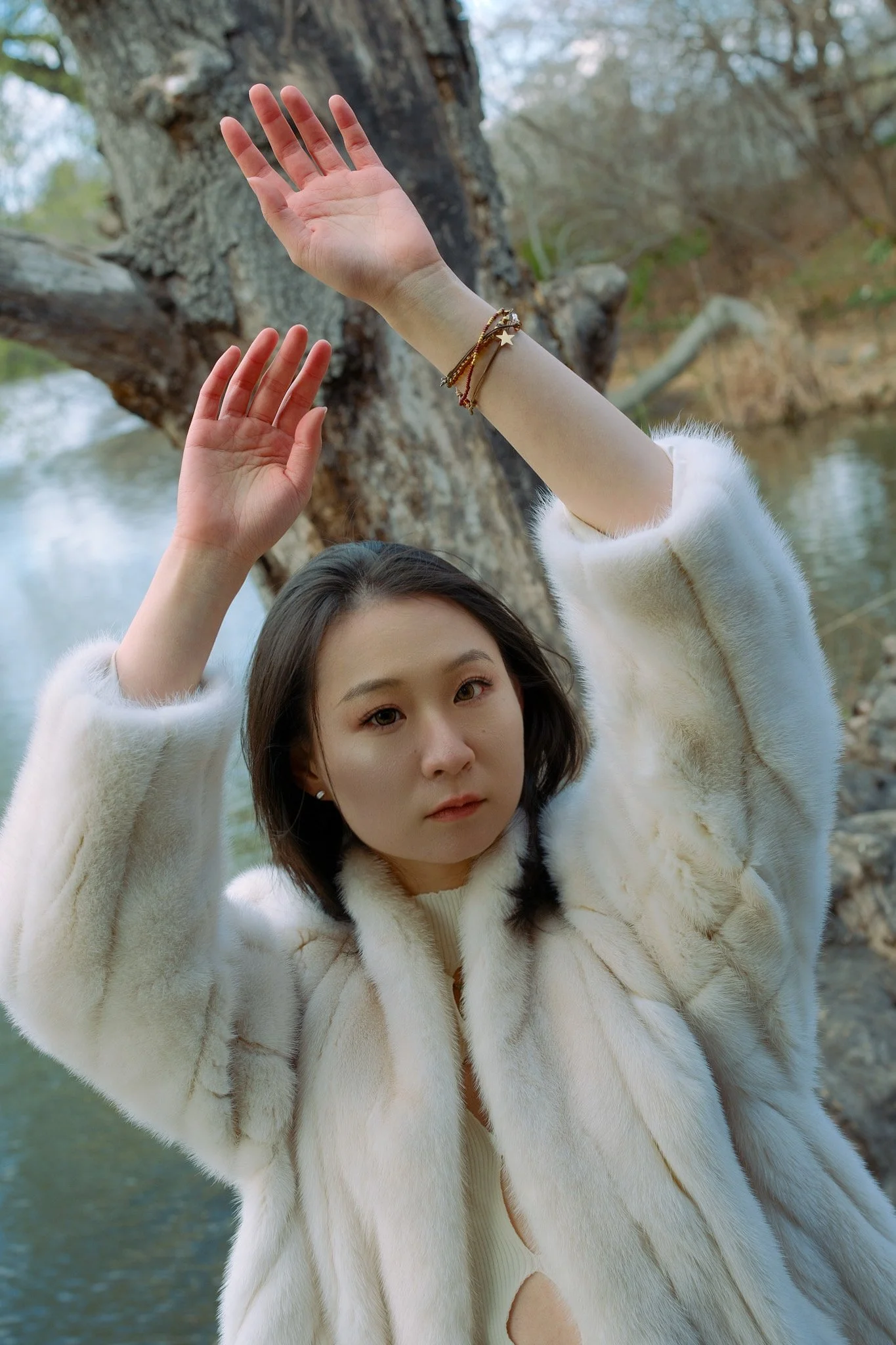 A woman in a white fur coat and bracelets stands near a tree and body of water, with her hands raised above her head as if posing or stretching.