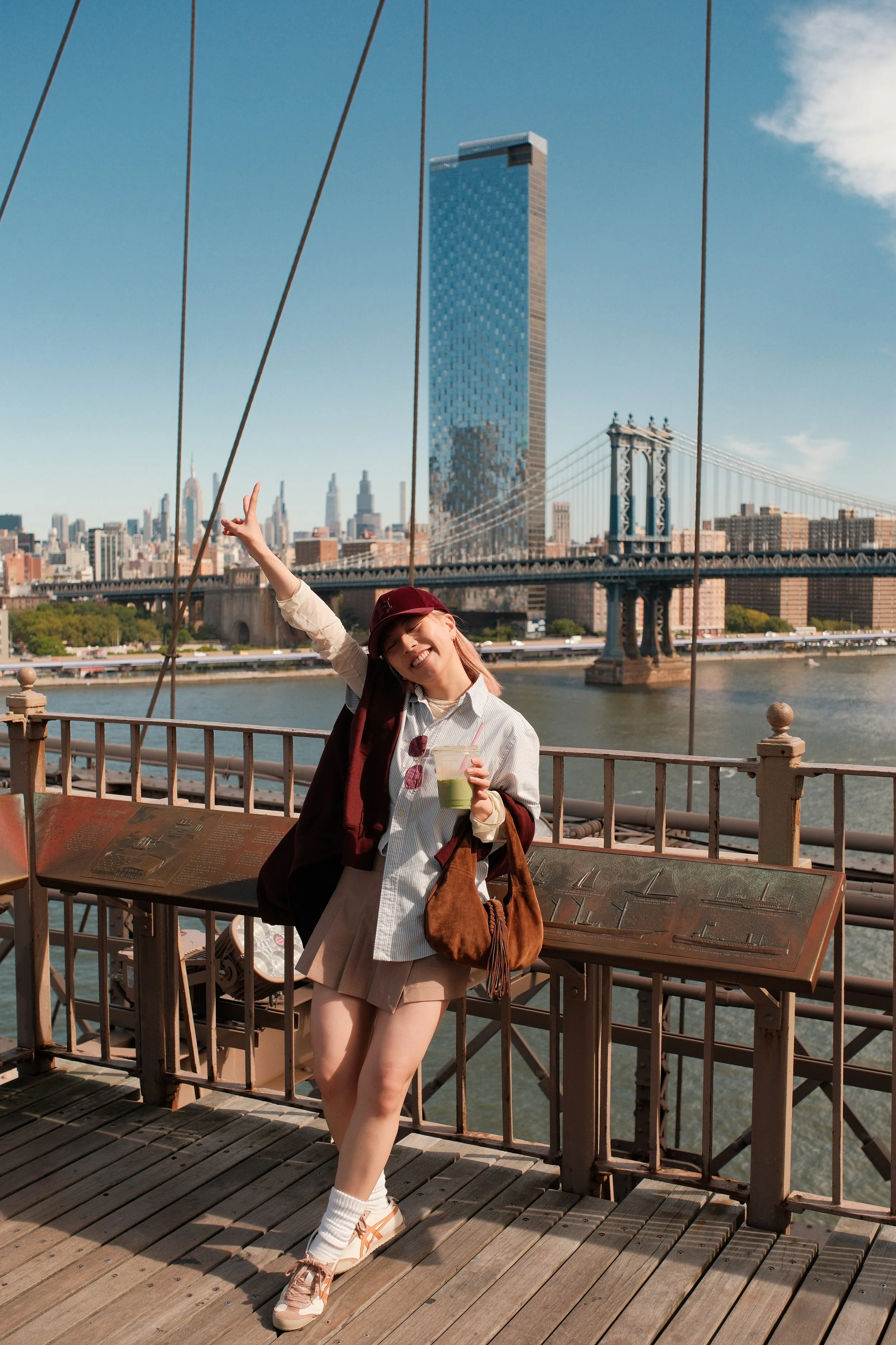 A young woman smiling and posing on the Brooklyn Bridge in New York City. She is holding a drink in her left hand, wearing a cap, sunglasses, a light striped shirt, a beige skirt, and sneakers. The city skyline, including the Manhattan Bridge and the