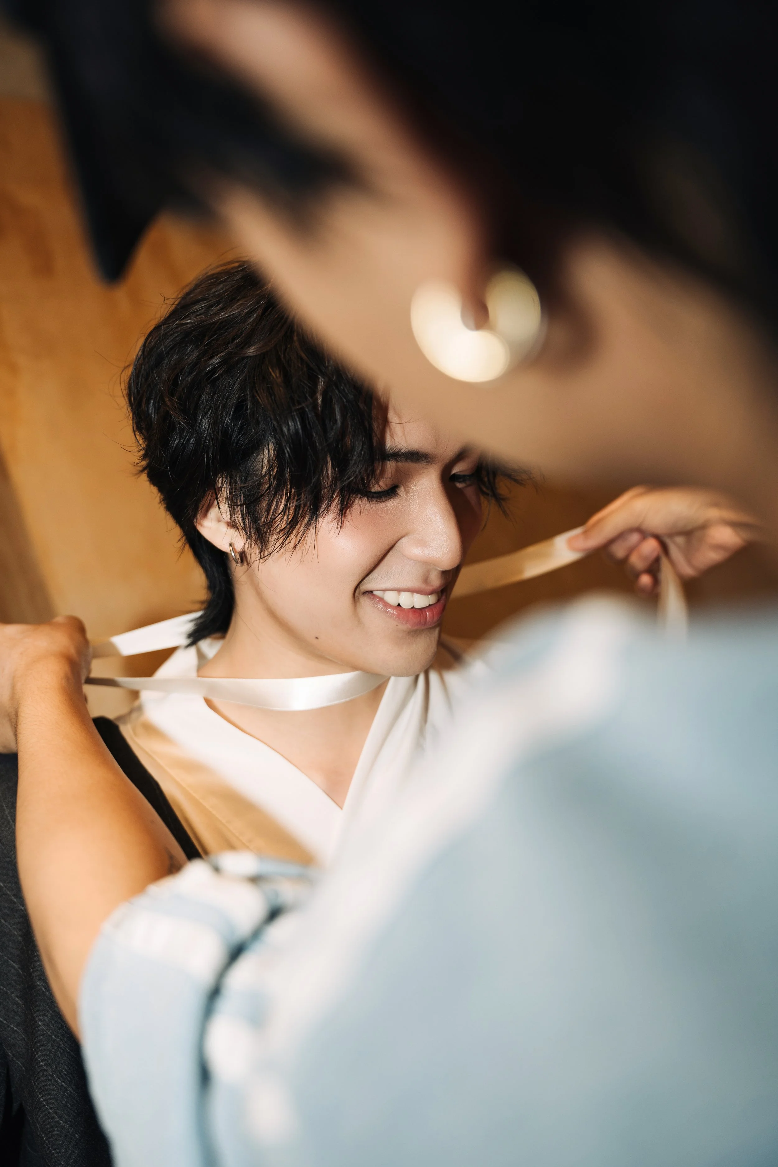 A person with short dark hair smiling while someone ties a white ribbon around their neck, with a warm indoor setting in the background.