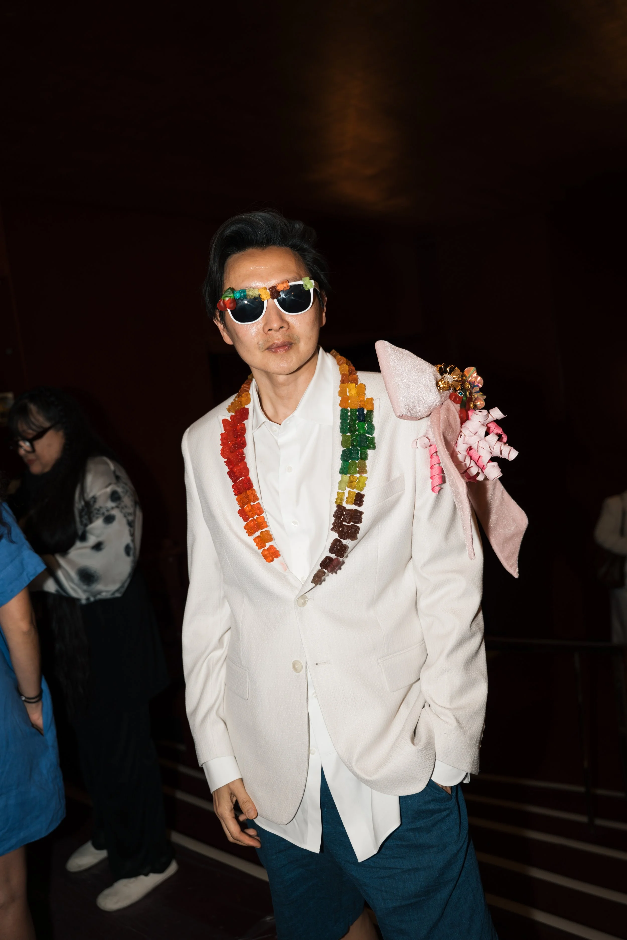 Person wearing sunglasses and a white suit jacket with colorful candy necklaces, posing at an indoor event.