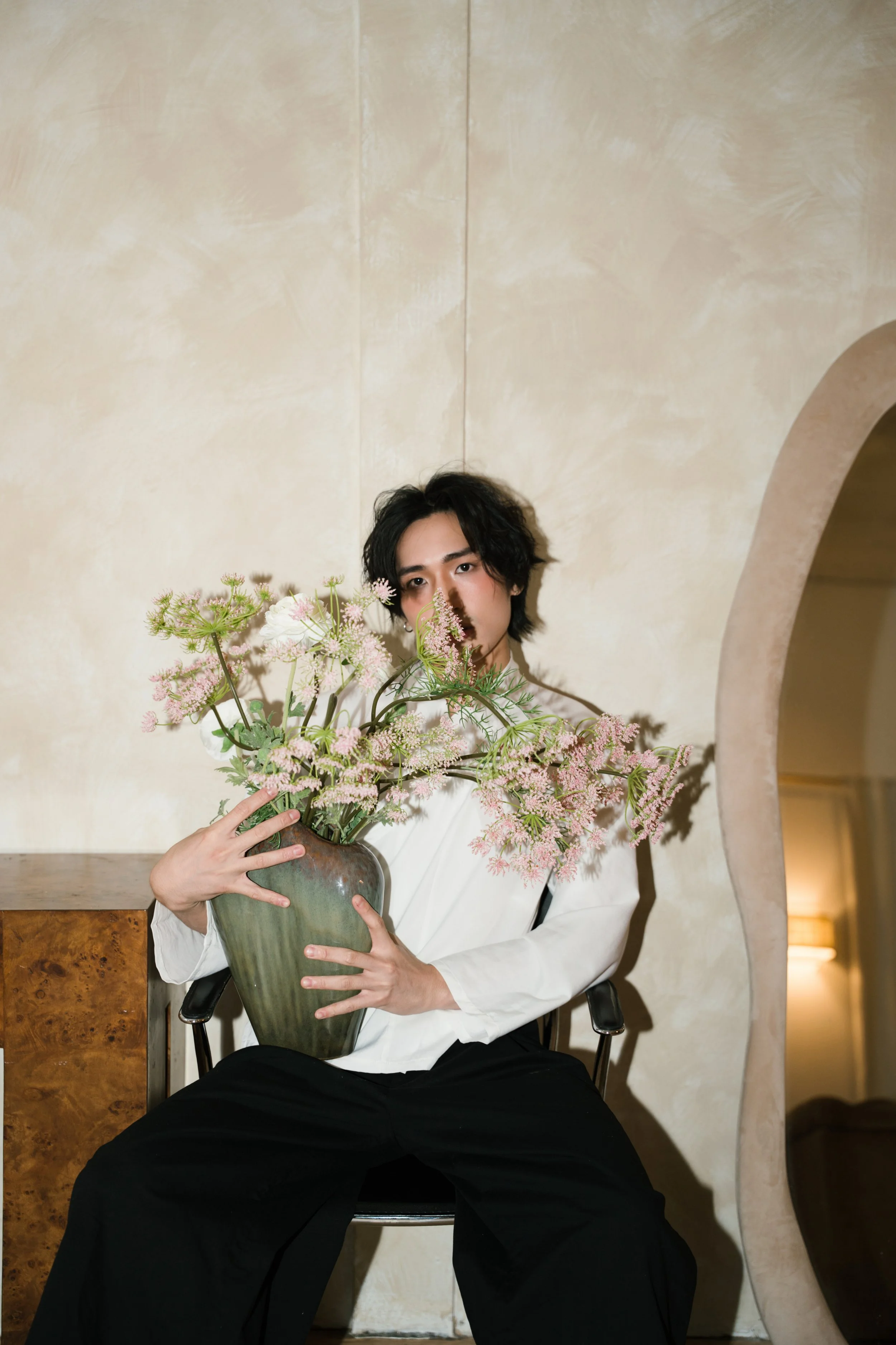 A person with dark, wavy hair sitting in a black chair, holding a large green vase filled with pink and white flowers, in a room with beige walls and an arched doorway.