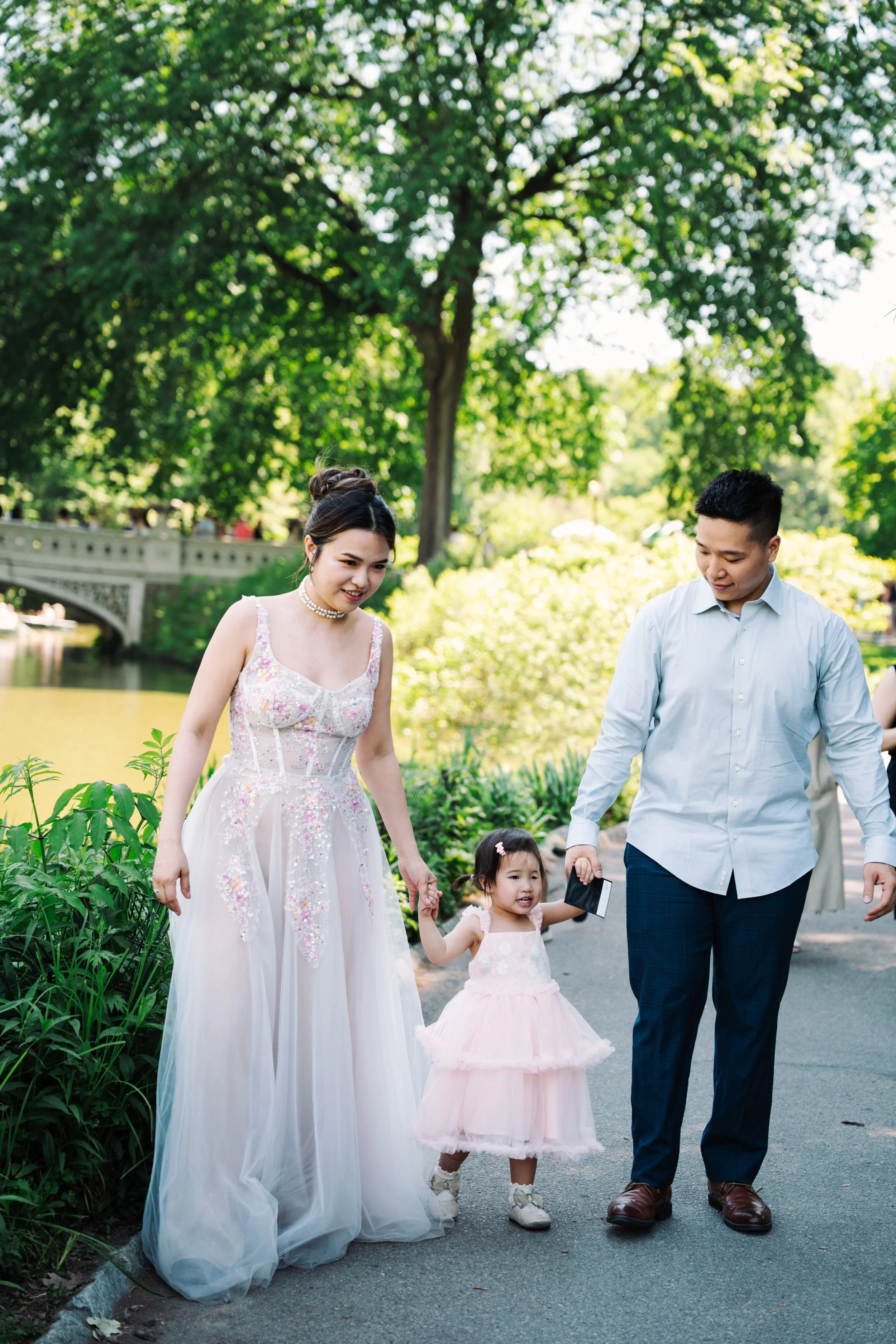 A woman in a white wedding dress and a man in a white shirt walk with a young girl in a pink dress through a park with lush green trees.