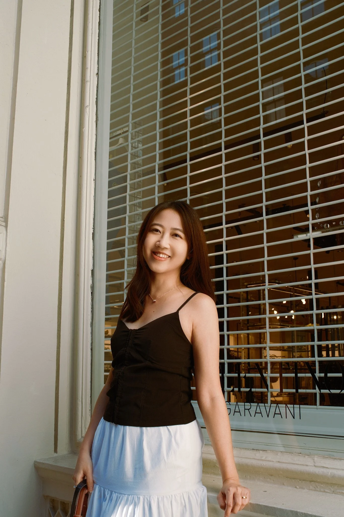 A young woman with long dark hair wearing a black sleeveless top and a white skirt standing outside in front of a window with a white grid and a glass storefront behind her. She is smiling and holding a small brown purse.