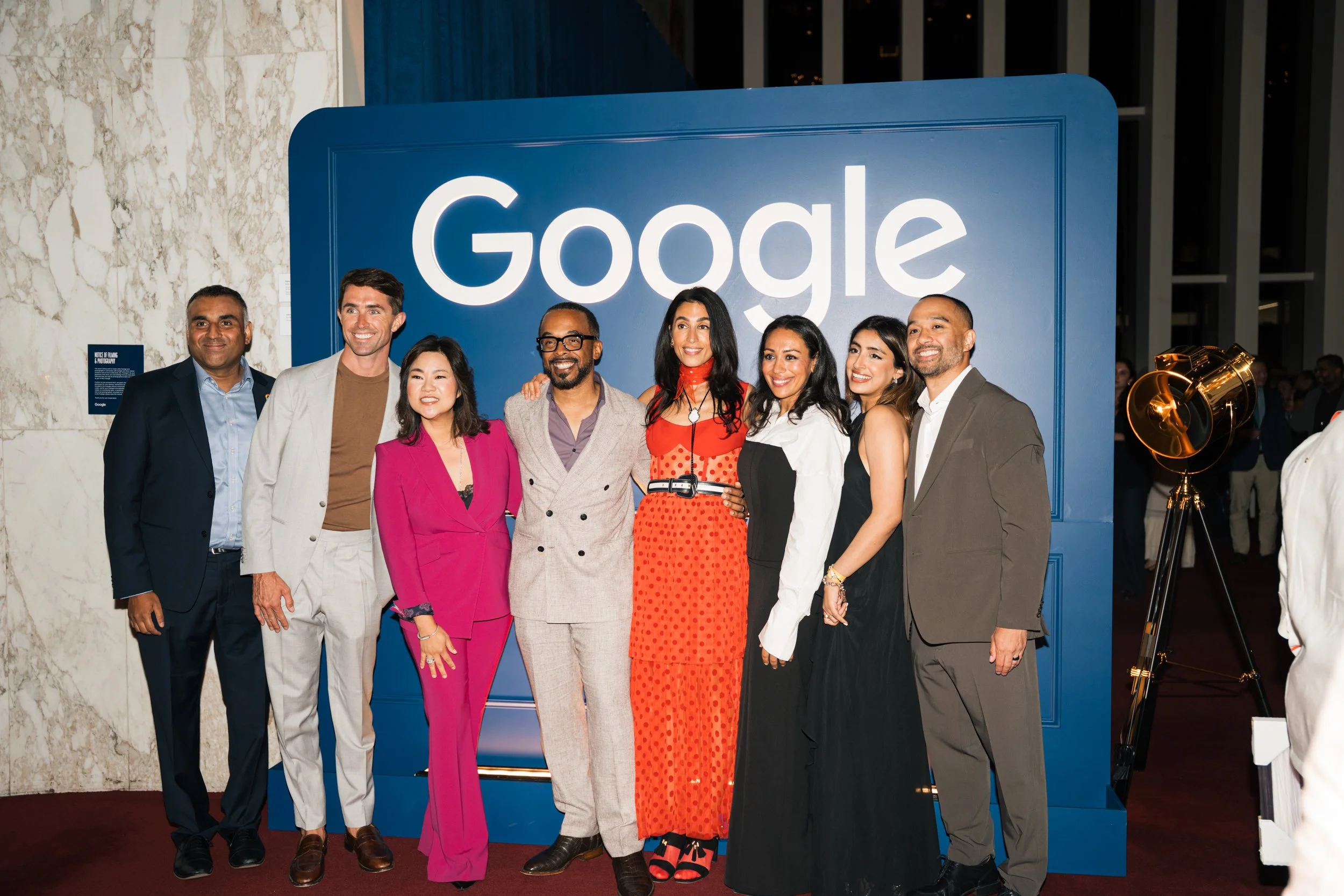 A group of nine diverse people standing together, posing in front of a large blue Google sign at an indoor event, smiling for the camera.