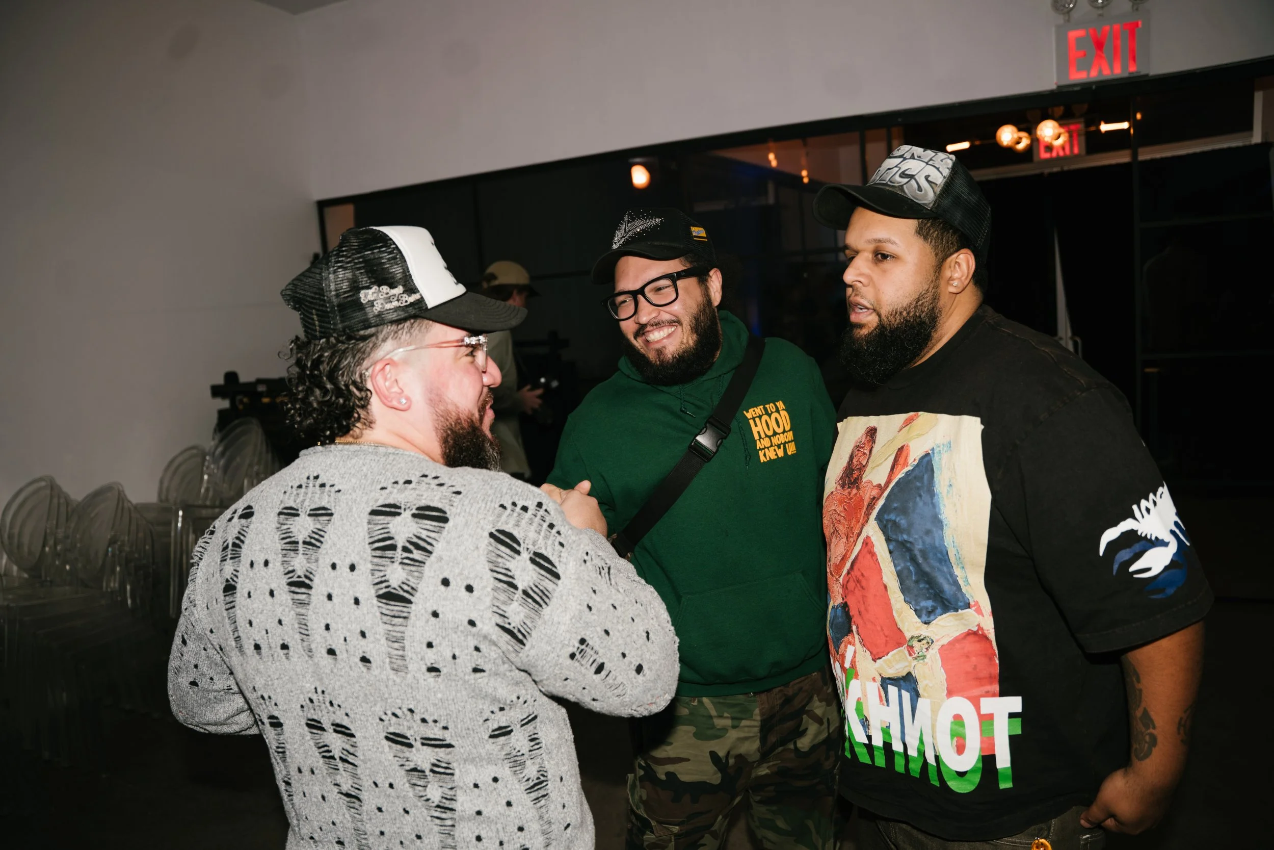 Three men engaged in conversation indoors, with chairs stacked against a wall, and an illuminated red 'EXIT' sign in the background.