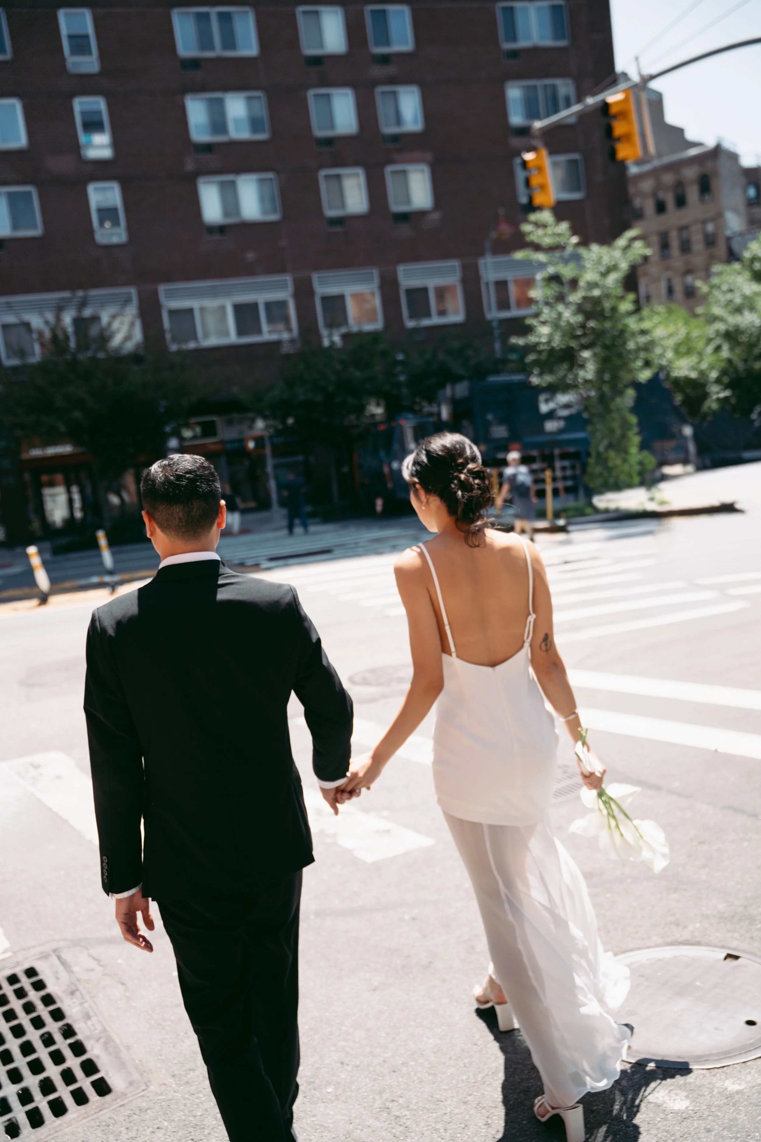 A bride and groom holding hands as they walk across a city street during daytime. The groom wears a black suit, and the bride wears a white dress and high heels, carrying a bouquet.