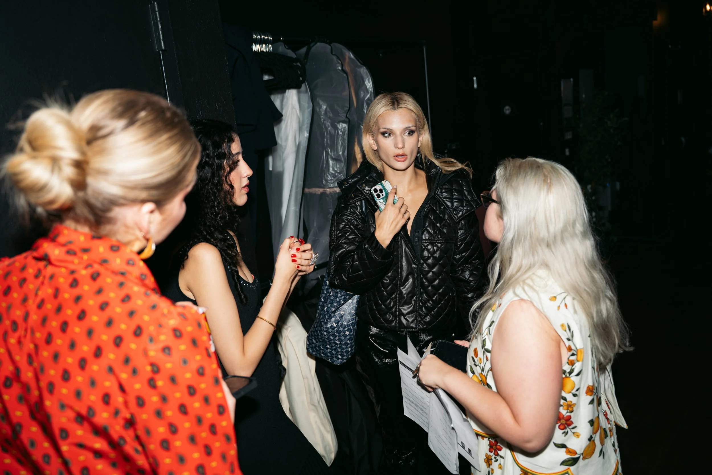 A blonde woman in a black quilted jacket speaking to a group of women in conversation backstage, with clothing on a rack in the background.