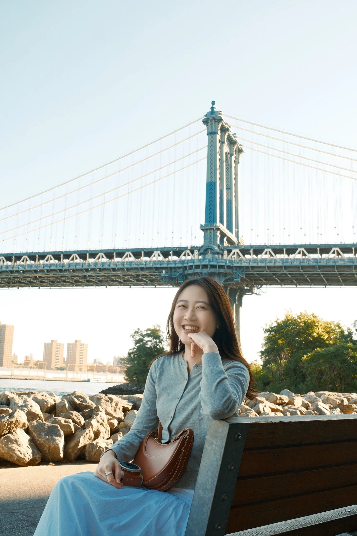 A woman sitting on a park bench near the water, smiling and holding a coffee cup, with the Manhattan Bridge in the background, trees, rocky shoreline, and city buildings.