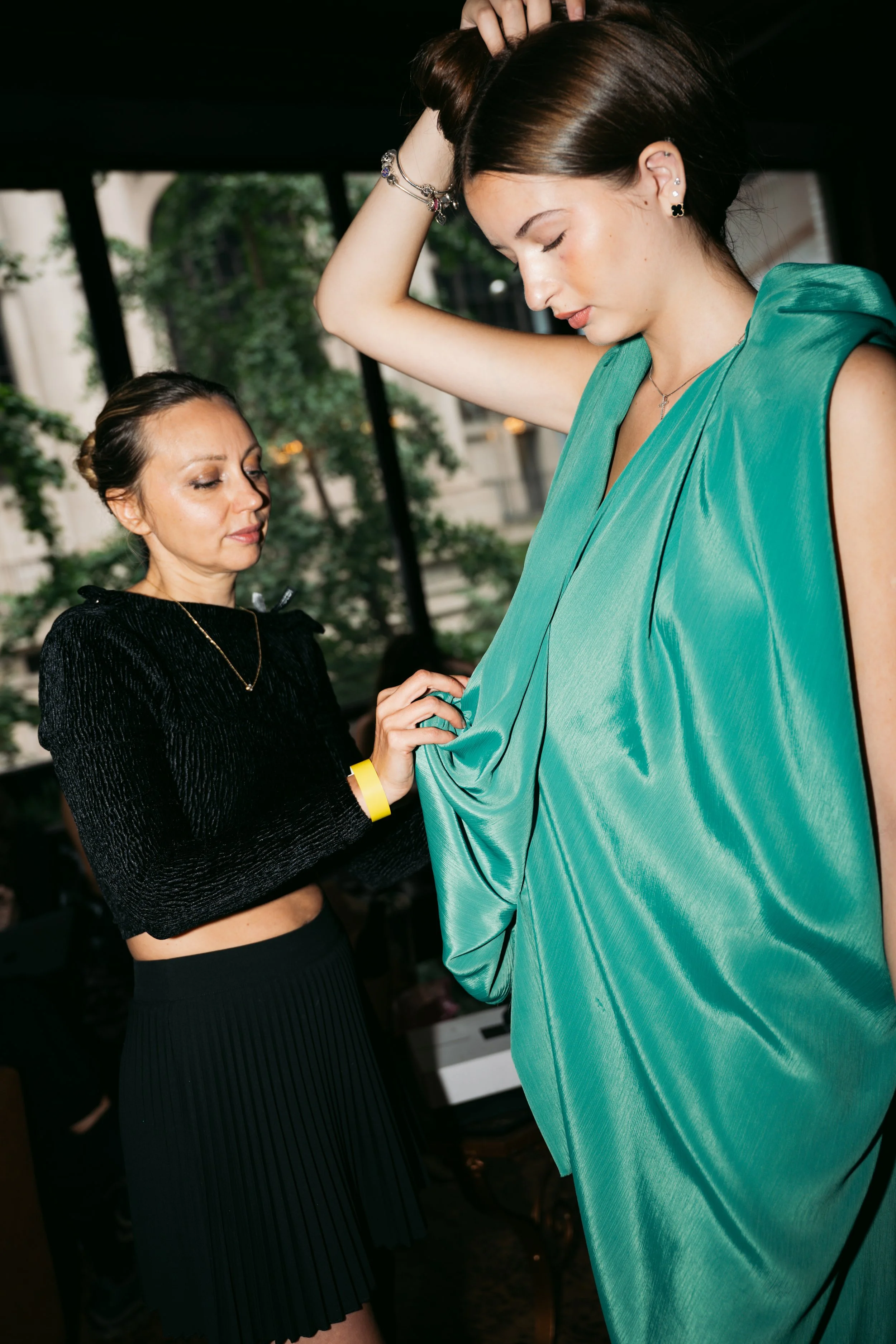 A woman in a green satin dress adjusting her hair, with a woman in a black outfit helping her with the dress in a brightly lit indoor setting.