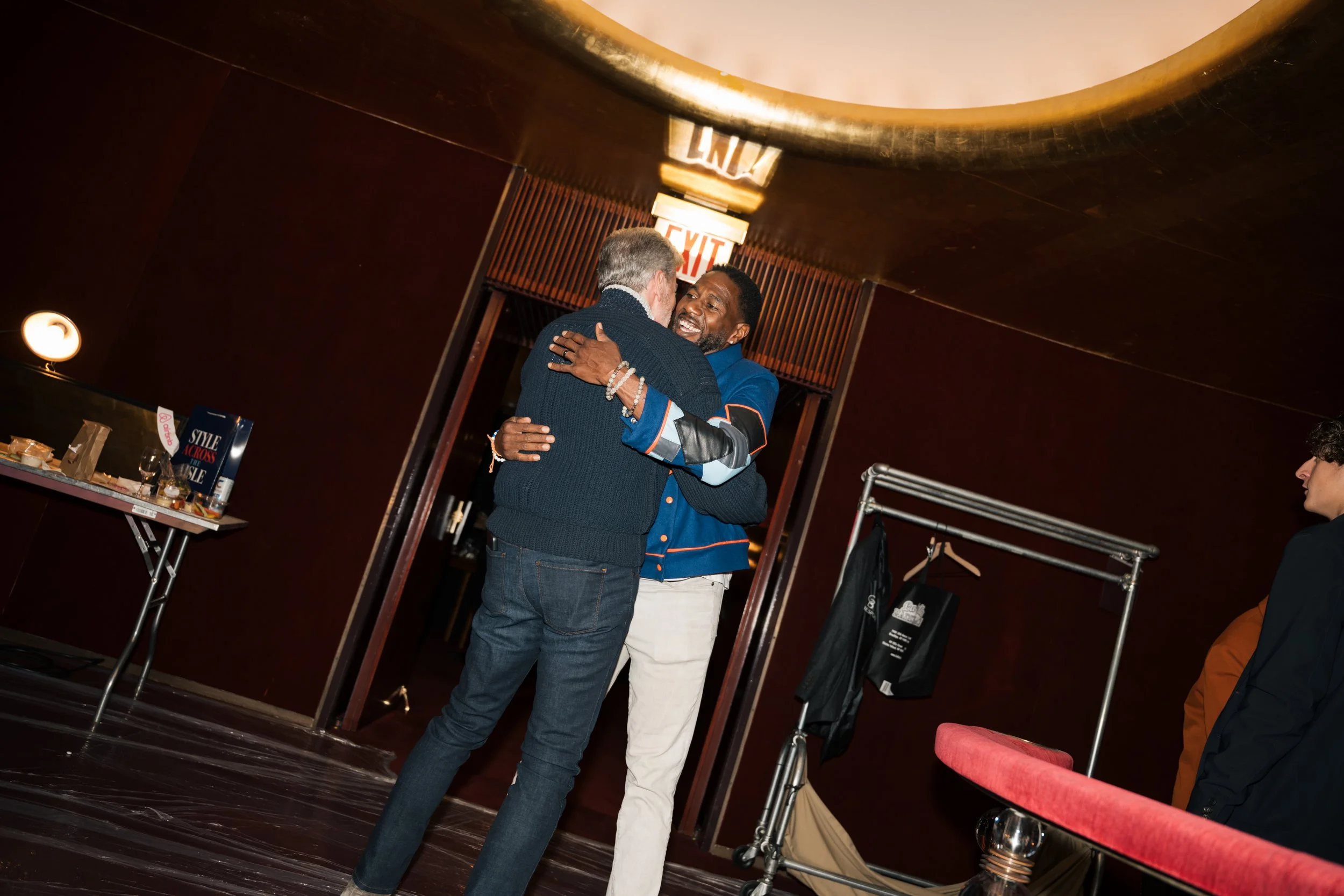 Two men hugging and smiling in a room with a dark ceiling, a sign in the background that reads 'EXIT,' and a table with food and magazines on it. One man has gray hair and a beard, wearing a dark sweater, and the other man has short black hair, weari