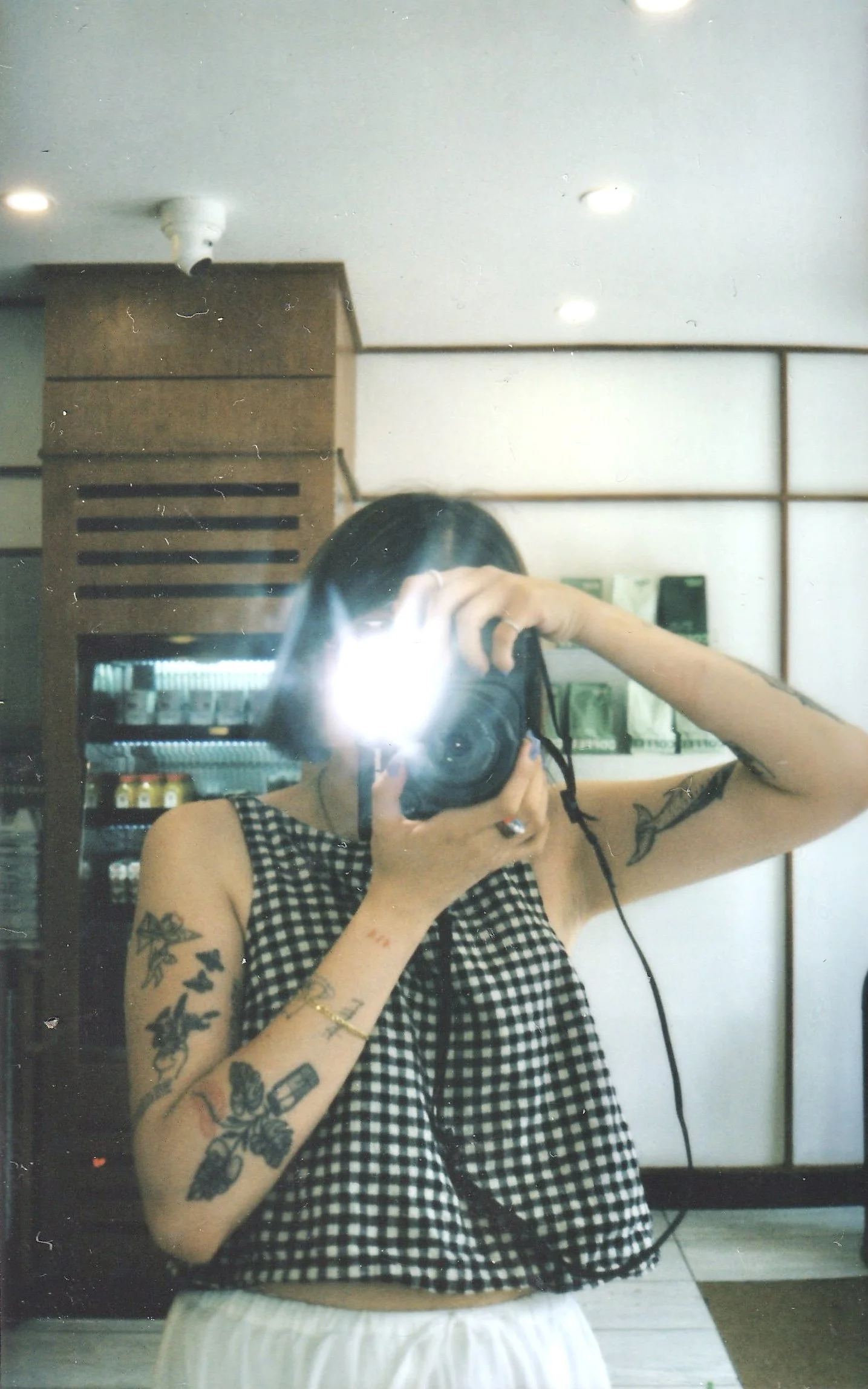 A person taking a mirror selfie with a camera, wearing a black and white checkered sleeveless top and white shorts. The person has tattoos on both arms and a ring on the left hand. The background features wooden and glass shelves, and a ceiling with recessed lighting.