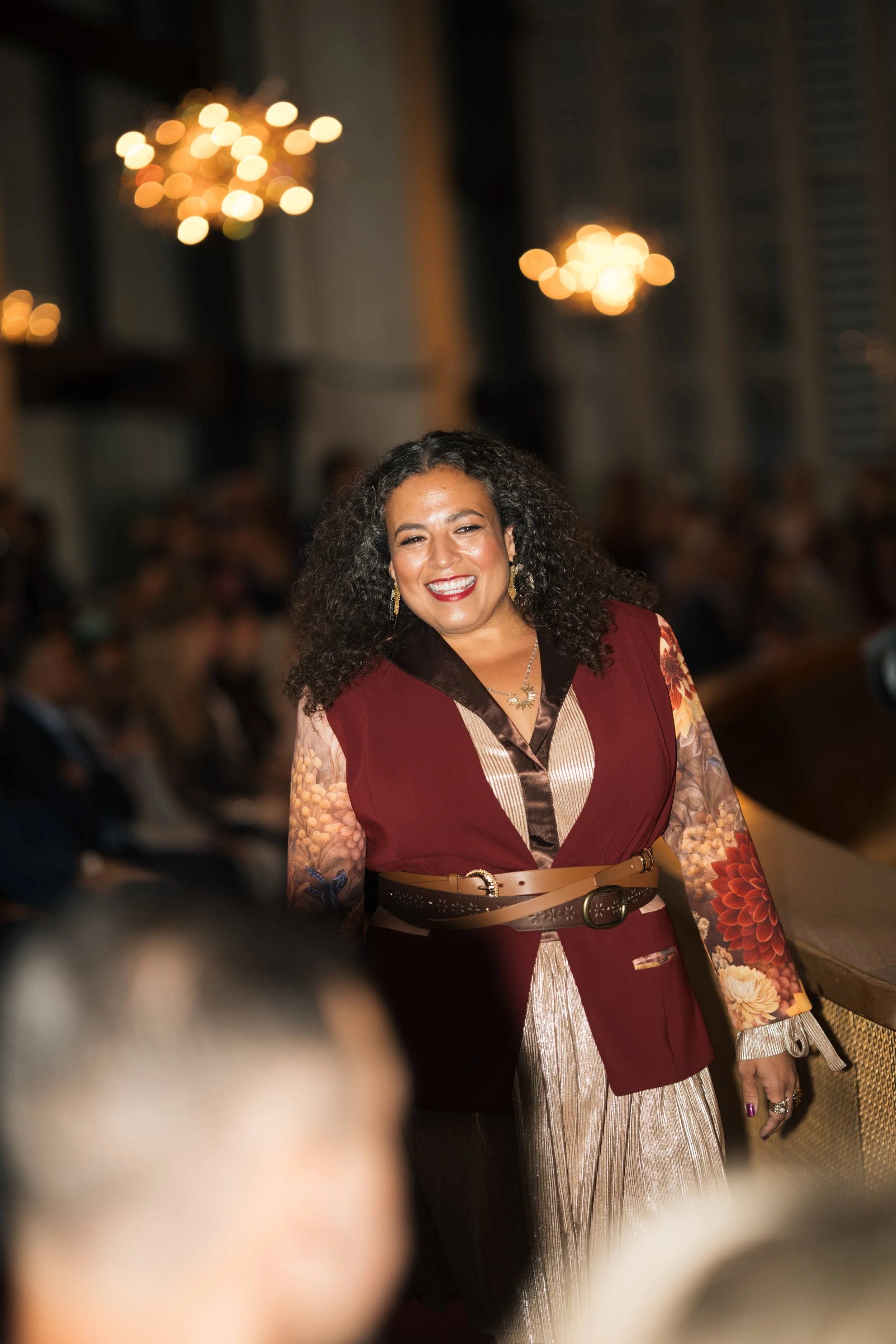 A woman with curly dark hair smiling at an indoor event with warm lighting and chandeliers in the background, wearing a patterned dress and a wide belt.