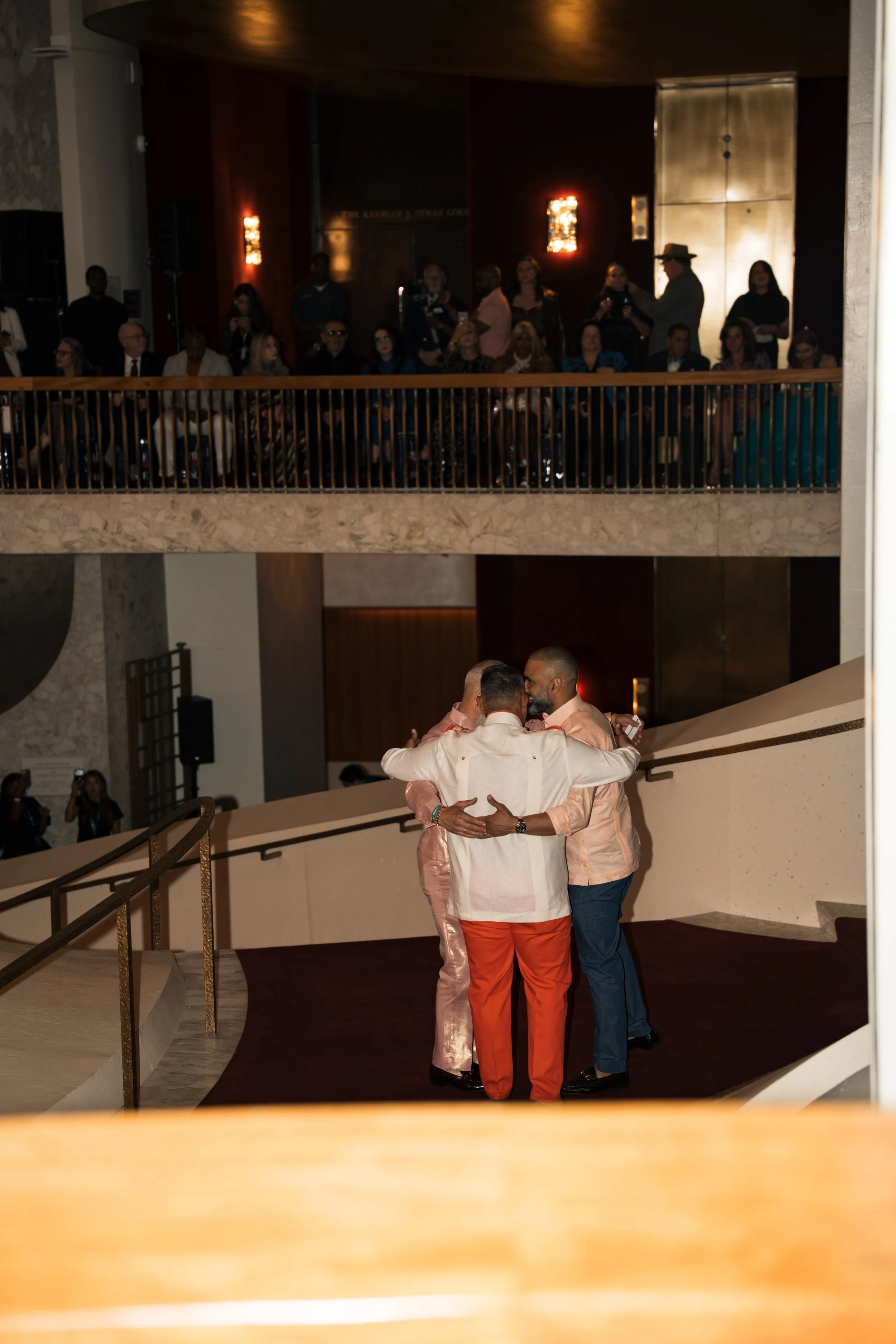 Two men embracing on a staircase, one wearing a peach shirt and the other wearing a white shirt with red pants, in a warmly lit indoor setting. In the background, a group of people are seated at a balcony overlooking the scene.