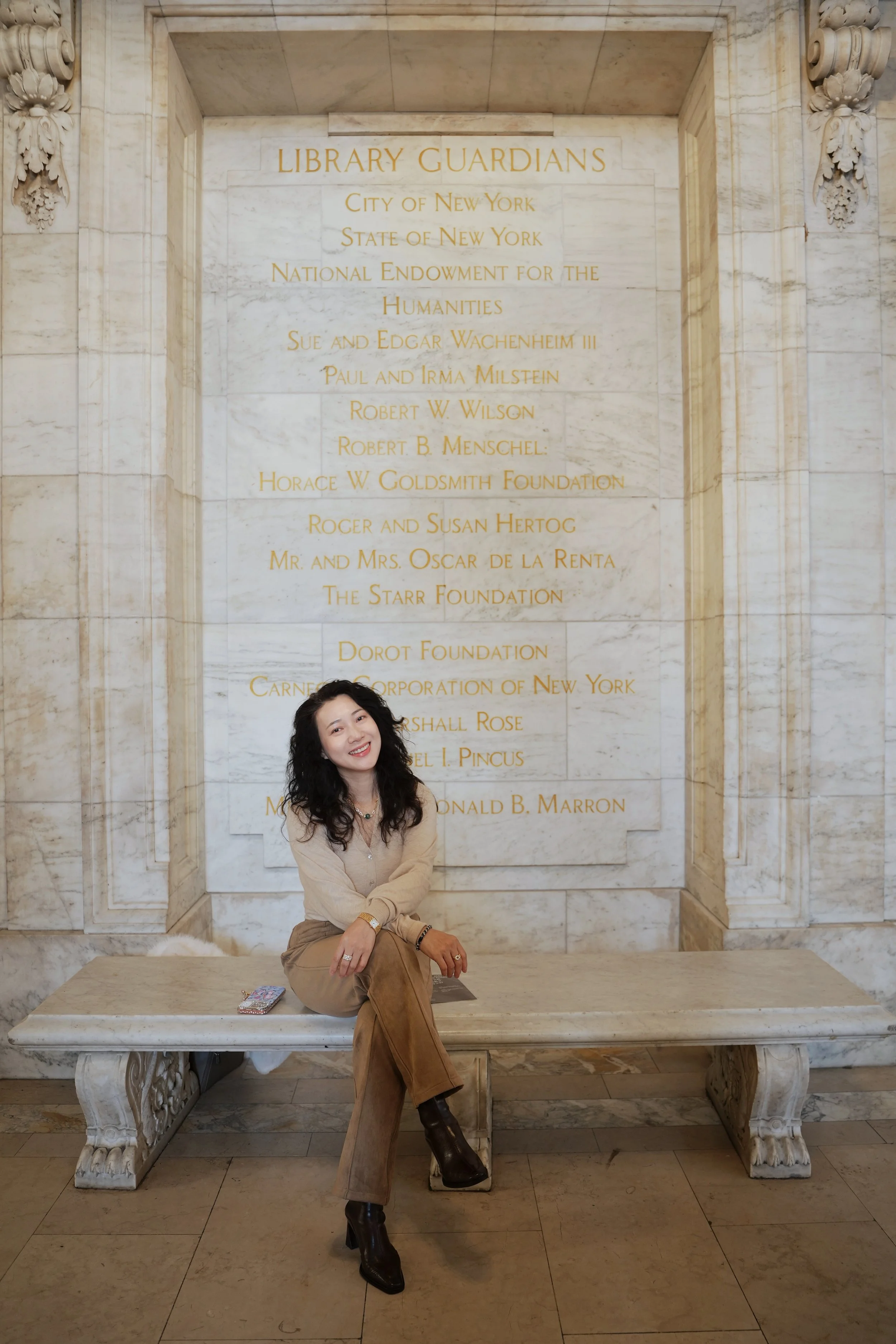 A woman with dark curly hair and wearing tan clothing, including a beige shirt, tan pants, and dark heeled boots, sitting on a marble bench inside a grand building with a carved stone wall behind her. The wall has gold-lettered inscriptions honoring 