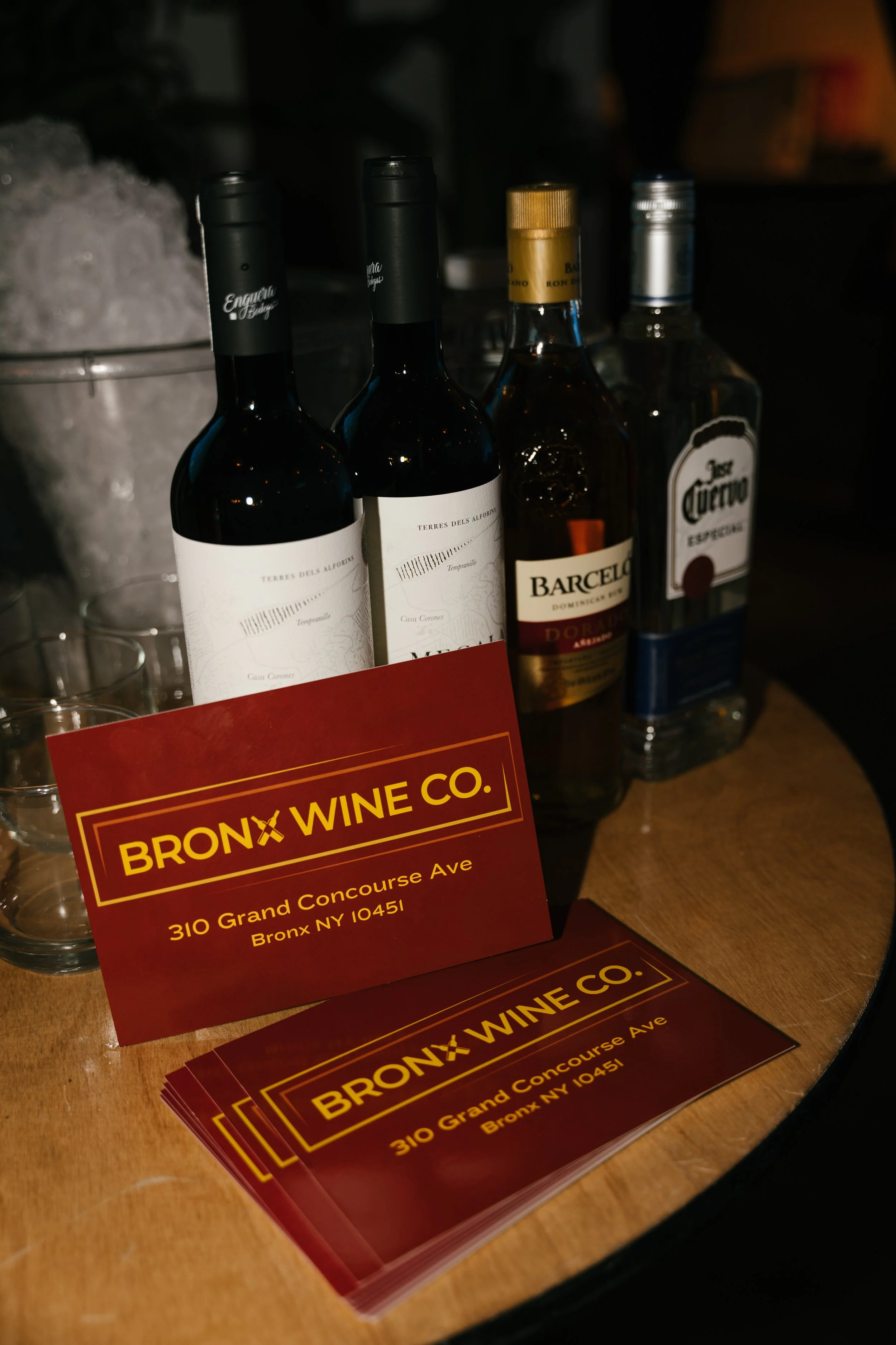 Stacks of red and yellow business cards for Bronx Wine Co. on a wooden table, with a partially visible bottle of alcohol and glasses in the background.