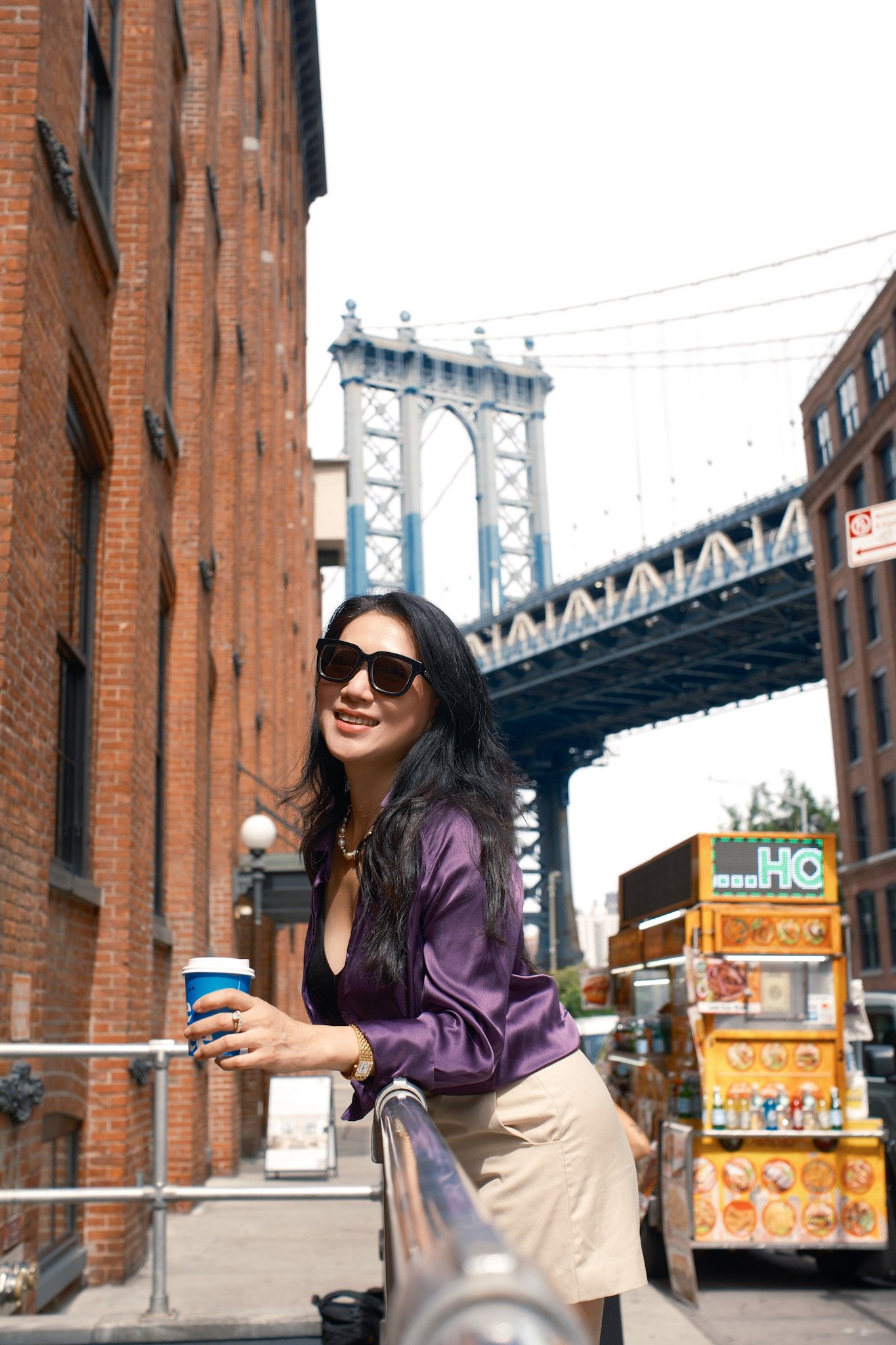 A woman with long black hair, wearing sunglasses, a purple satin jacket, and a beige skirt, holding a cup of coffee, standing on a city street near the Brooklyn Bridge.