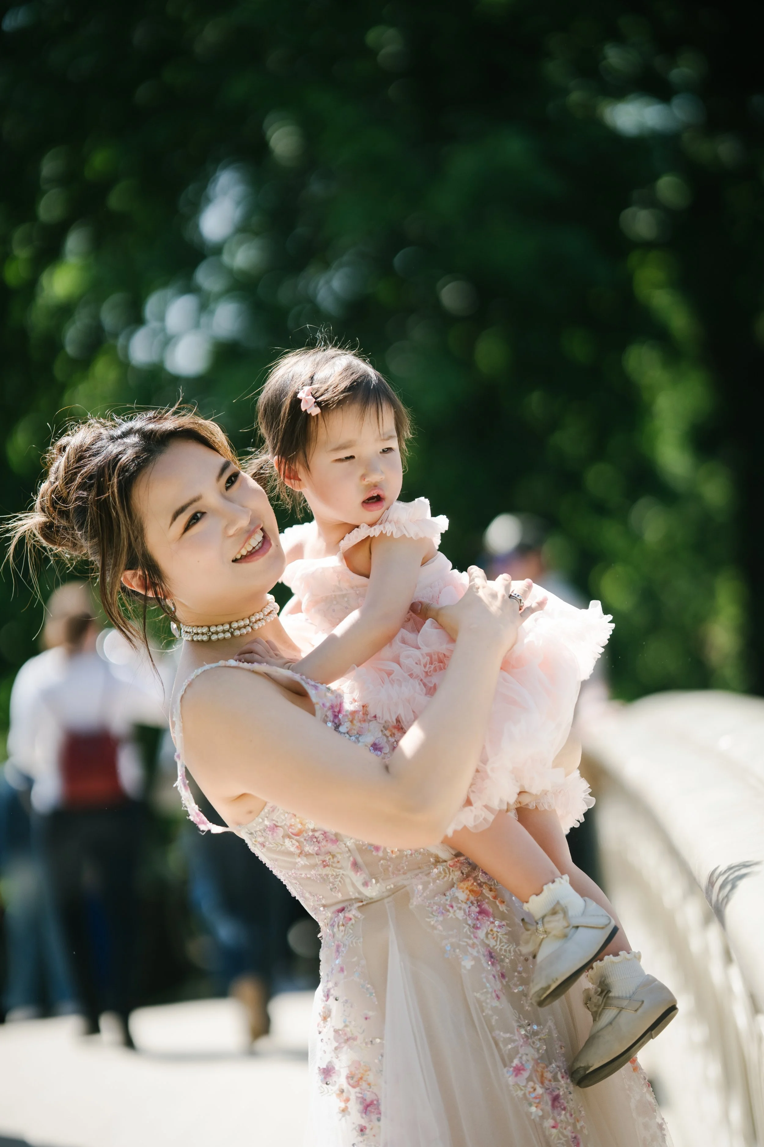 A woman with a pearl necklace smiling while holding a young girl in a pink dress with ruffles and floral accents outdoors in a park.
