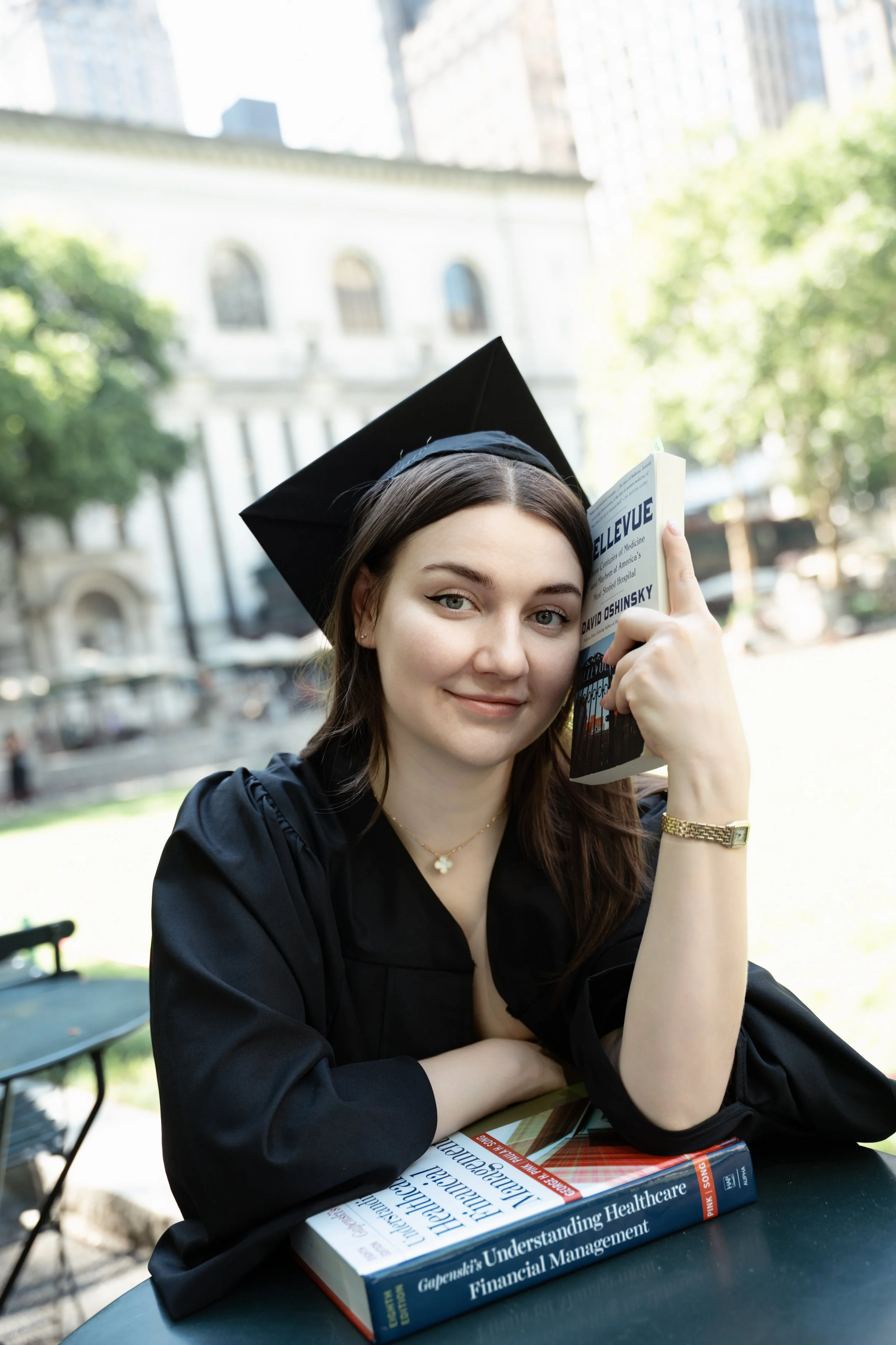 A young woman in a black graduation cap and gown sitting at an outdoor table, holding a book titled 'Gapecki's Understanding Healthcare Financial Management,' and smiling at the camera.