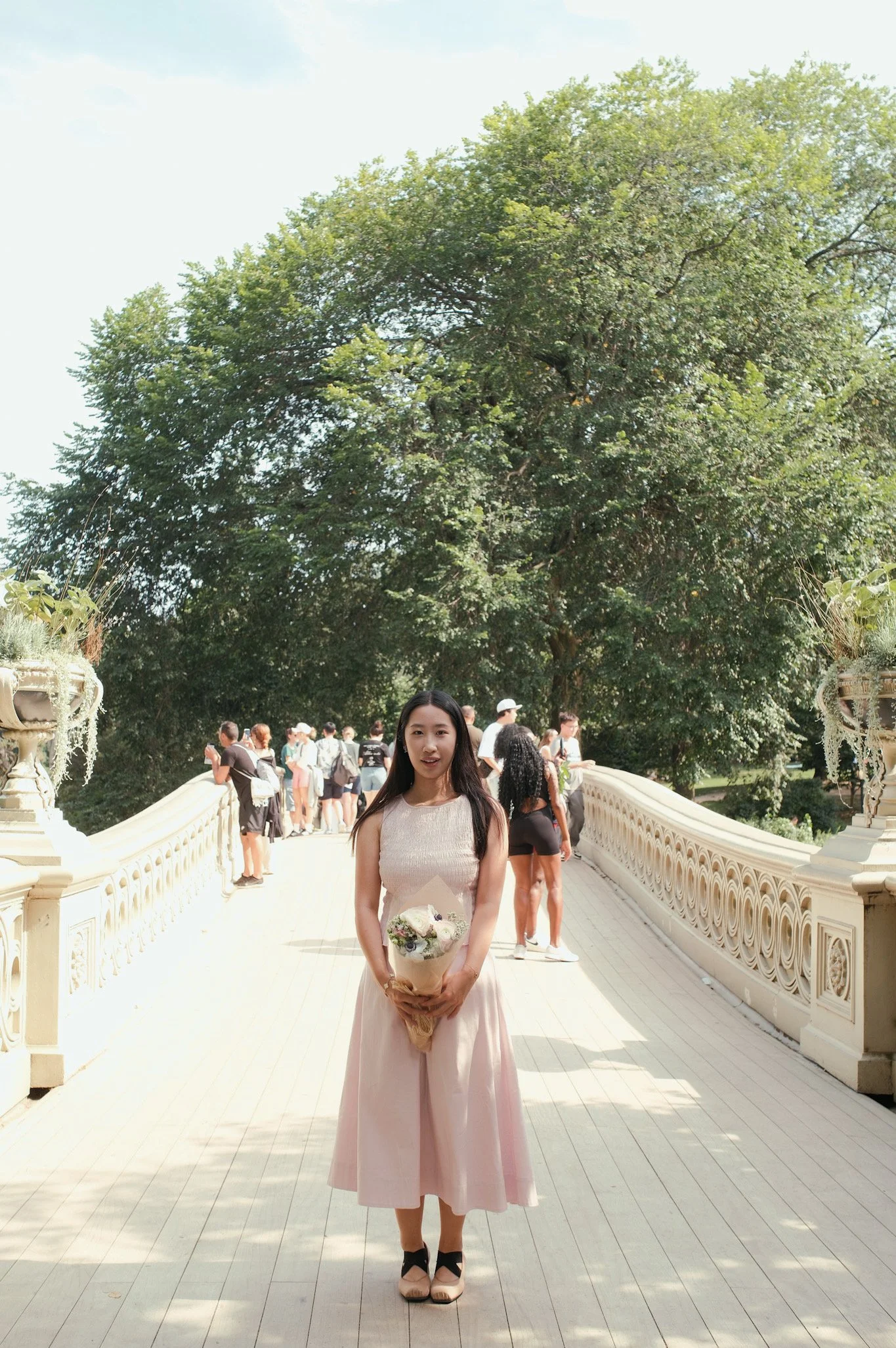 A woman in a light pink dress holding a bouquet of flowers, standing on a white bridge with a large tree behind her and other people in the background.