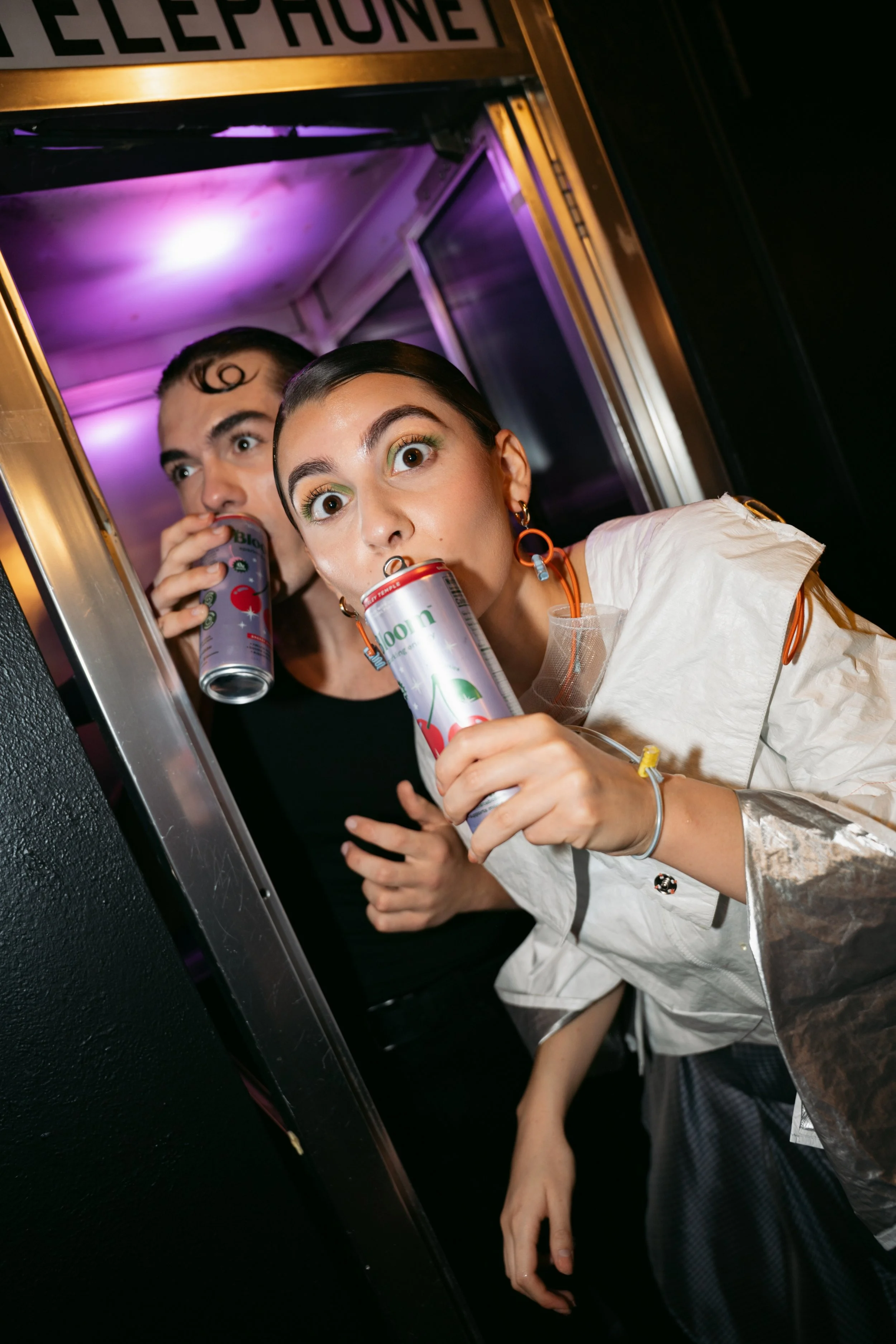Two people with colorful makeup and styled hair are leaning out of a food truck window, drinking soda cans, with a sign above the window that says 'TELEPHONE'.